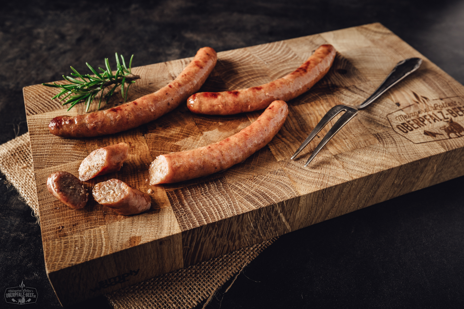 Vier rohe Salsiccia-Bratwürste auf Holzbrett – hochwertige italienische Grillwurst Vier grobe Wildbratwürste mit sichtbaren Kräutern und Gewürzen liegen nebeneinander auf einer dunklen Schieferplatte, begleitet von Rosmarinzweigen – hochwertige, rustikale Wildwurst aus der Oberpfalz, ideal für Grillfans und Wildfleischliebhaber.