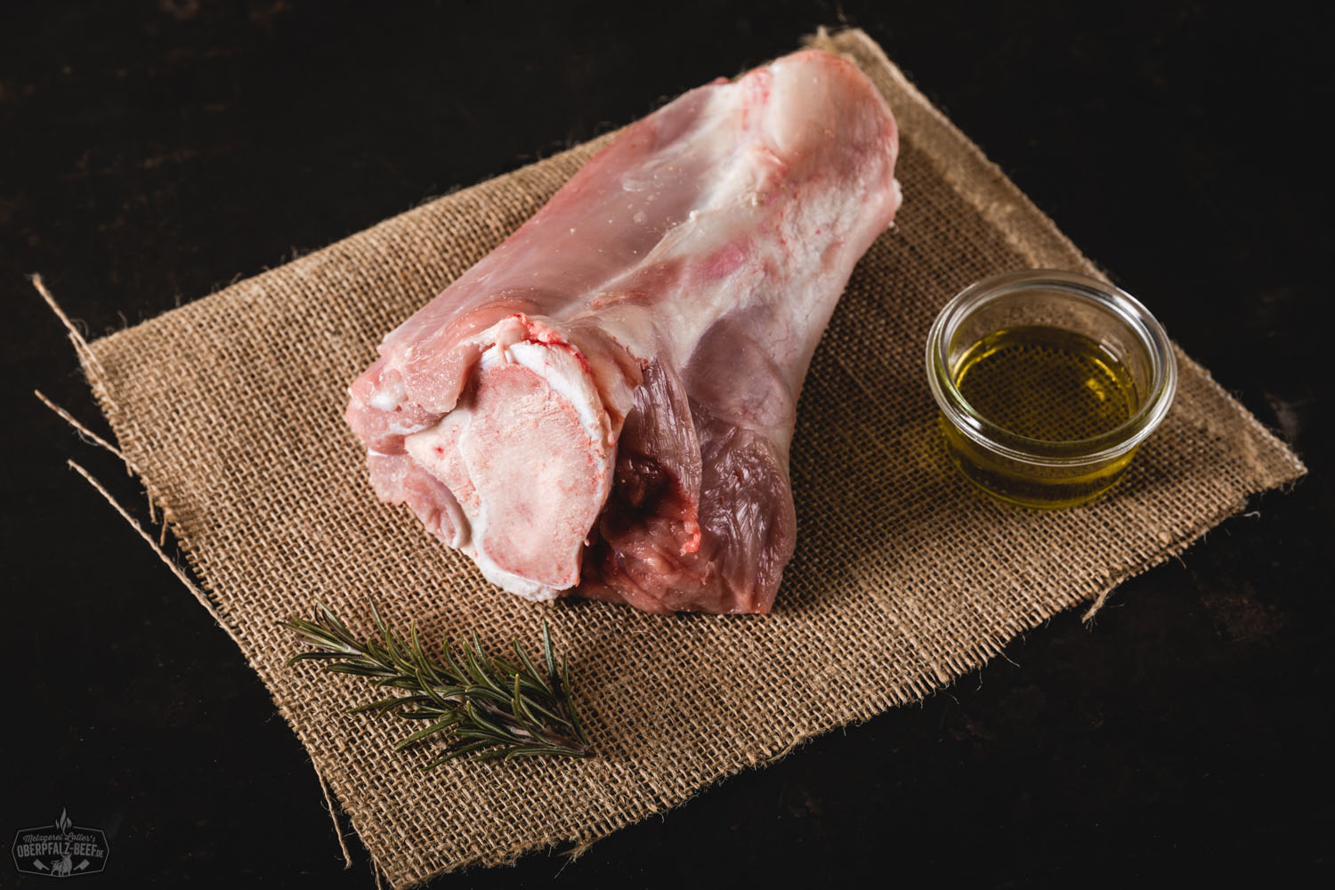 Rohe hintere Kalbshaxe vom Oberpfälzer Milchkalb auf dunkler Schieferplatte, umgeben von Rosmarinzweigen, Knoblauchzehen, Meersalz und buntem Pfeffer – hochwertiges Kalbfleisch für Schmorgerichte, Metzgerqualität aus der Oberpfalz.