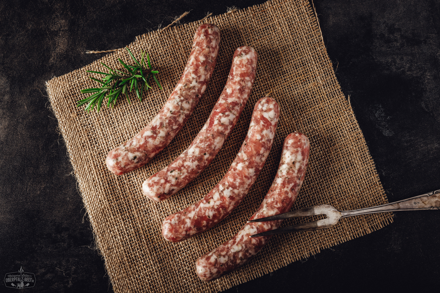 Vier frische Wildbratwürste aus regionaler Herstellung auf Holzbrett Bild zeigt vier stilvoll arrangierte Wildbratwürste aus der Oberpfalz auf einem rustikalen Holztisch, umgeben von aromatischen Kräutern, die die Qualität und Tradition regionaler Fleischspezialitäten unterstreichen.