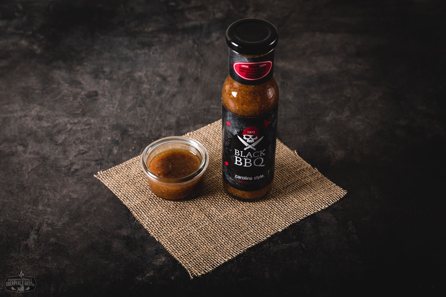 Black BBQ Sauce Carolina Style – Raucharomatische Grillsoße im amerikanischen Stil Flasche Black BBQ Sauce im Carolina Style von Oberpfalz Beef vor weißem Hintergrund, mit schwarzem Etikett, orangefarbenen Akzenten und stilisiertem Rindskopf-Logo – rauchige BBQ-Sauce im US-Südstaatenstil.