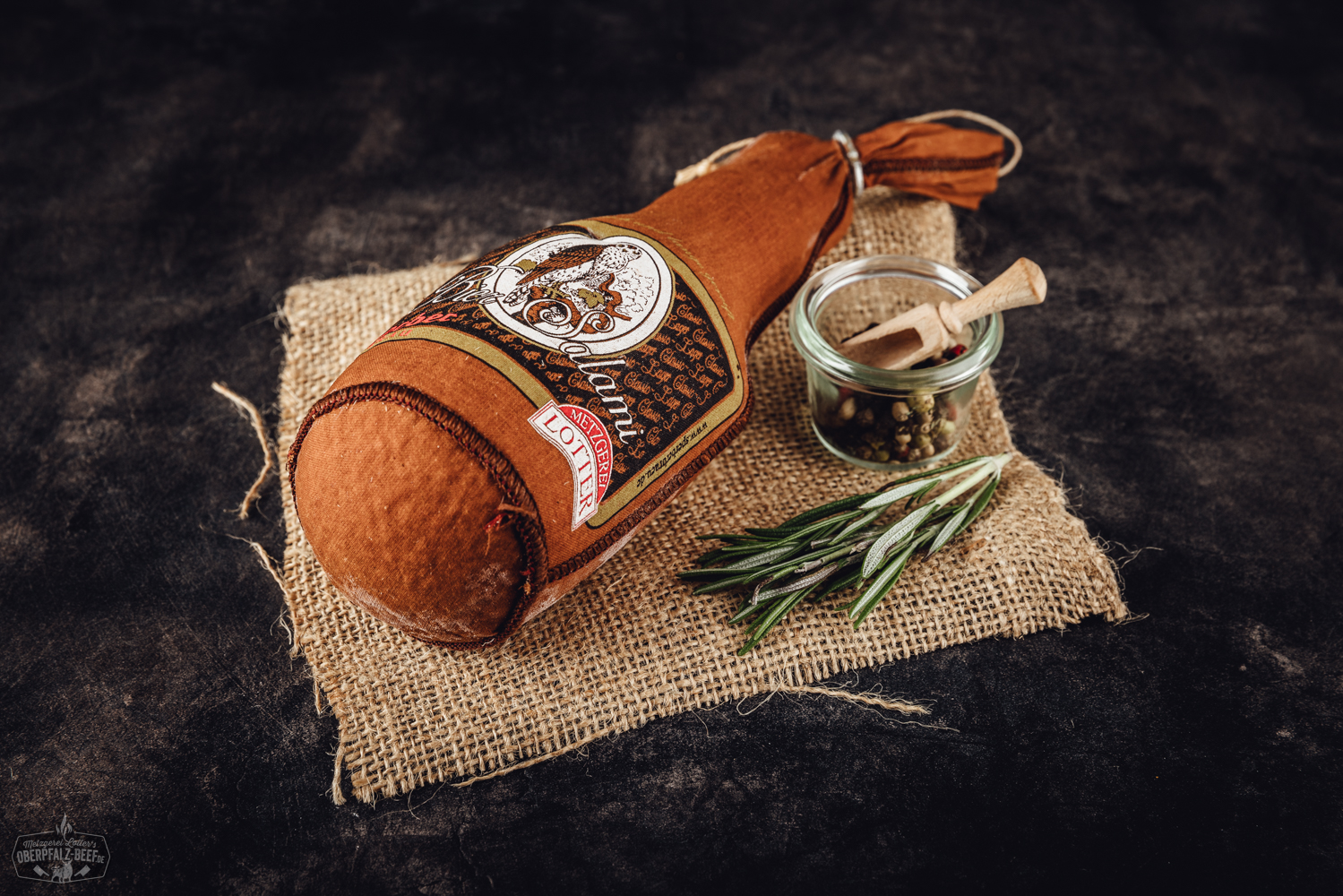 Nahaufnahme einer Biersalami auf einem Holzbrett mit angeschnittener Scheibe, umgeben von Pfefferkörnern und einem Bierglas im Hintergrund – hochwertige Delikatesswurst aus der Oberpfalz.