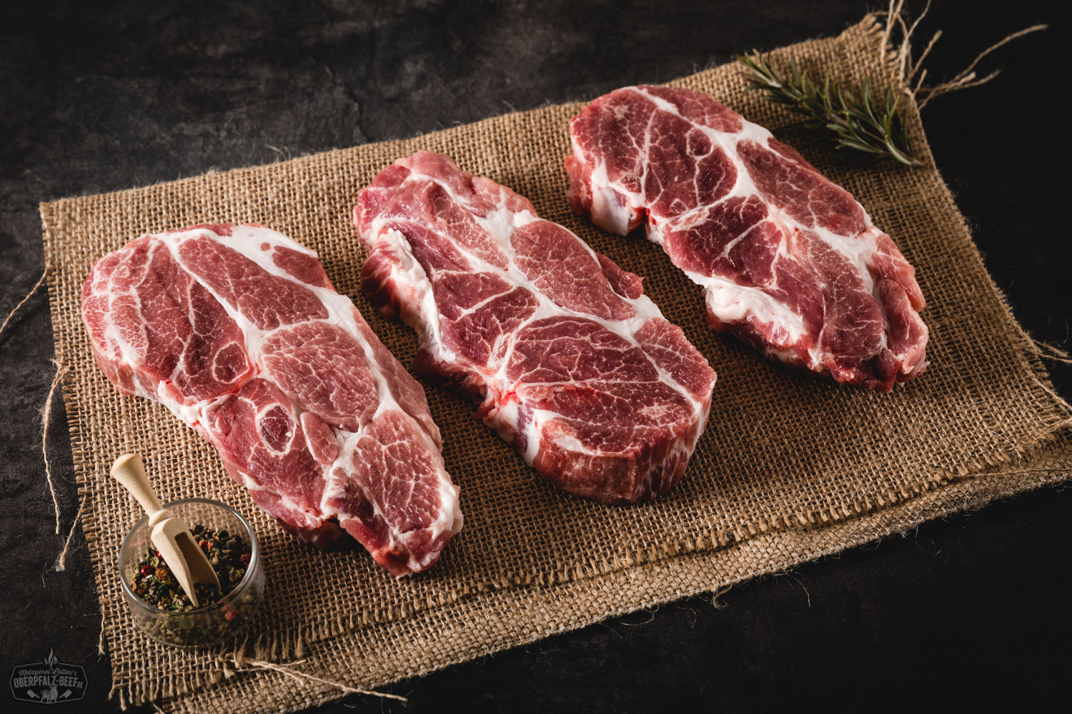 Zwei rohe Halssteaks vom Oberpfälzer Landschwein liegen nebeneinander auf rustikalem Holzbrett, umgeben von frischen Kräutern, Knoblauchzehen, Pfefferkörnern und Rosmarinzweigen – naturbelassen, frisch und bereit zum Grillen oder Braten.