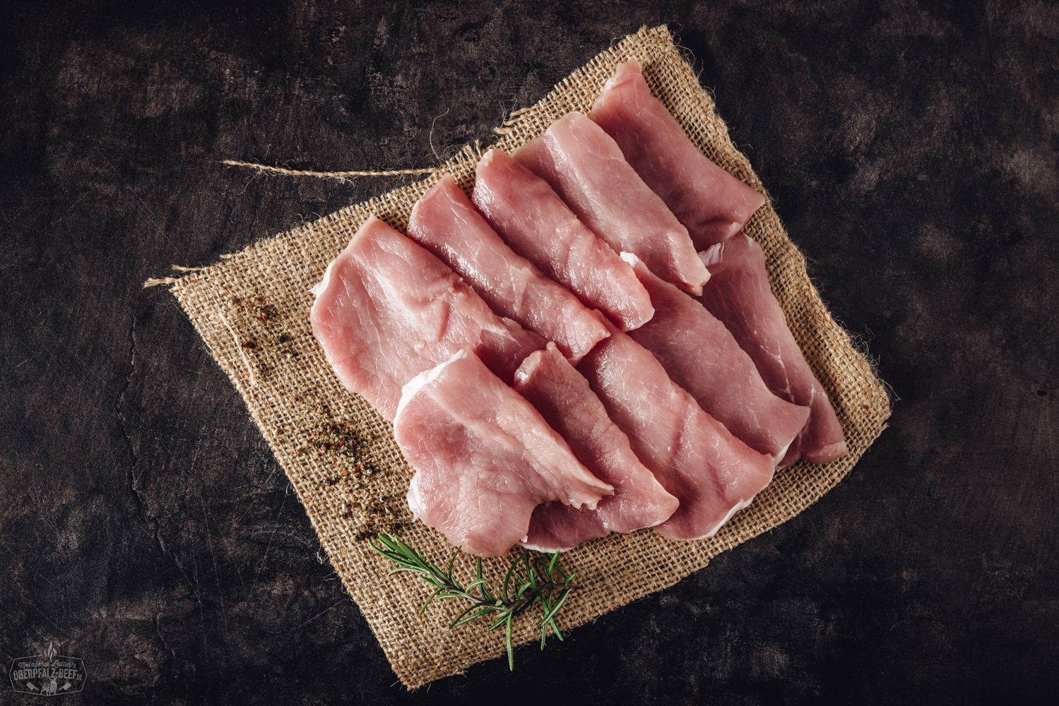 Handgeschnittenes Raclette-Fleisch vom Schweinerücken – hochwertiges Grillfleisch für Raclette und Fondue Handgeschnittene Raclette-Fleischscheiben aus dem Schweinerücken, fein marmoriert und portionsweise auf einem dunklen Holzbrett angerichtet, im Hintergrund Kräuter und Gewürze – hochwertige Zubereitungsidee für Raclette oder Grillgerichte.