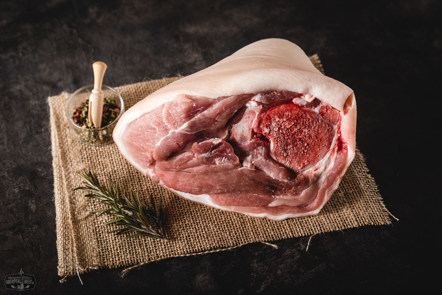 Hintere Haxe vom Oberpfälzer Landschwein – Regionales Schweinefleisch kaufen Rohe hintere Schweinshaxe vom Oberpfälzer Landschwein auf dunkler Steinplatte, umgeben von Rosmarinzweigen, Knoblauchzehen und buntem Pfeffer – ideal zum Schmoren oder Grillen.