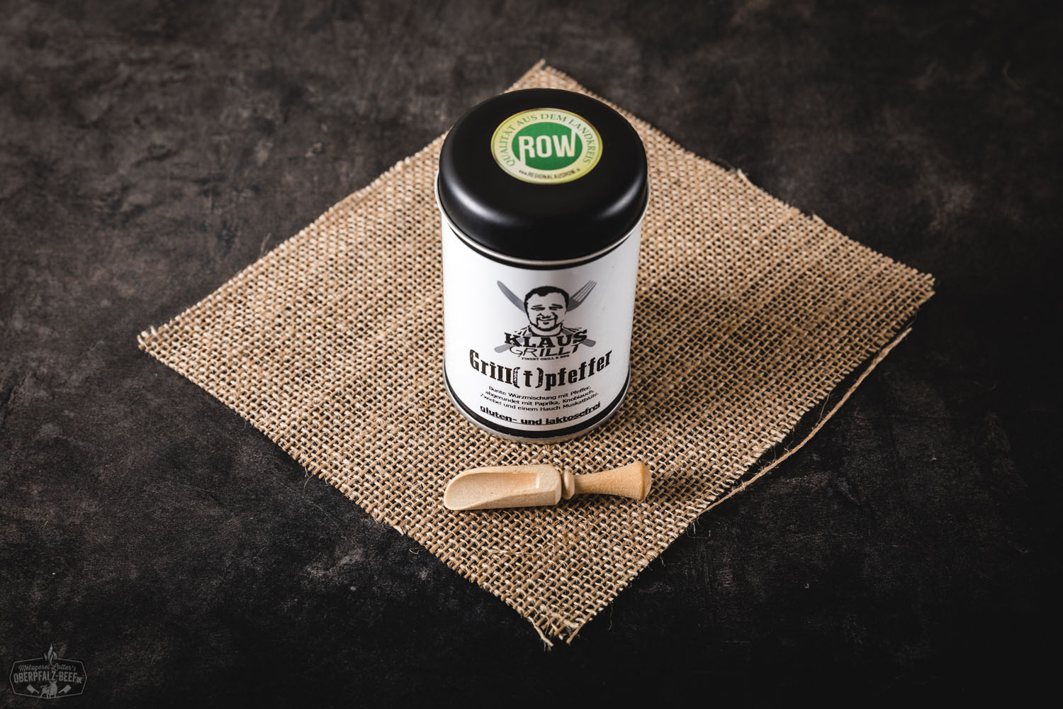 Mann steht an Grillstation im Freien und hält schwarzen Pfefferstreuer über aufgeschnittene Steaks auf Holzbrett, im Hintergrund rauchender Gasgrill und Holzfass, sommerliche Atmosphäre, Fokus auf Gewürzvorgang beim Grillen von Rindfleisch.