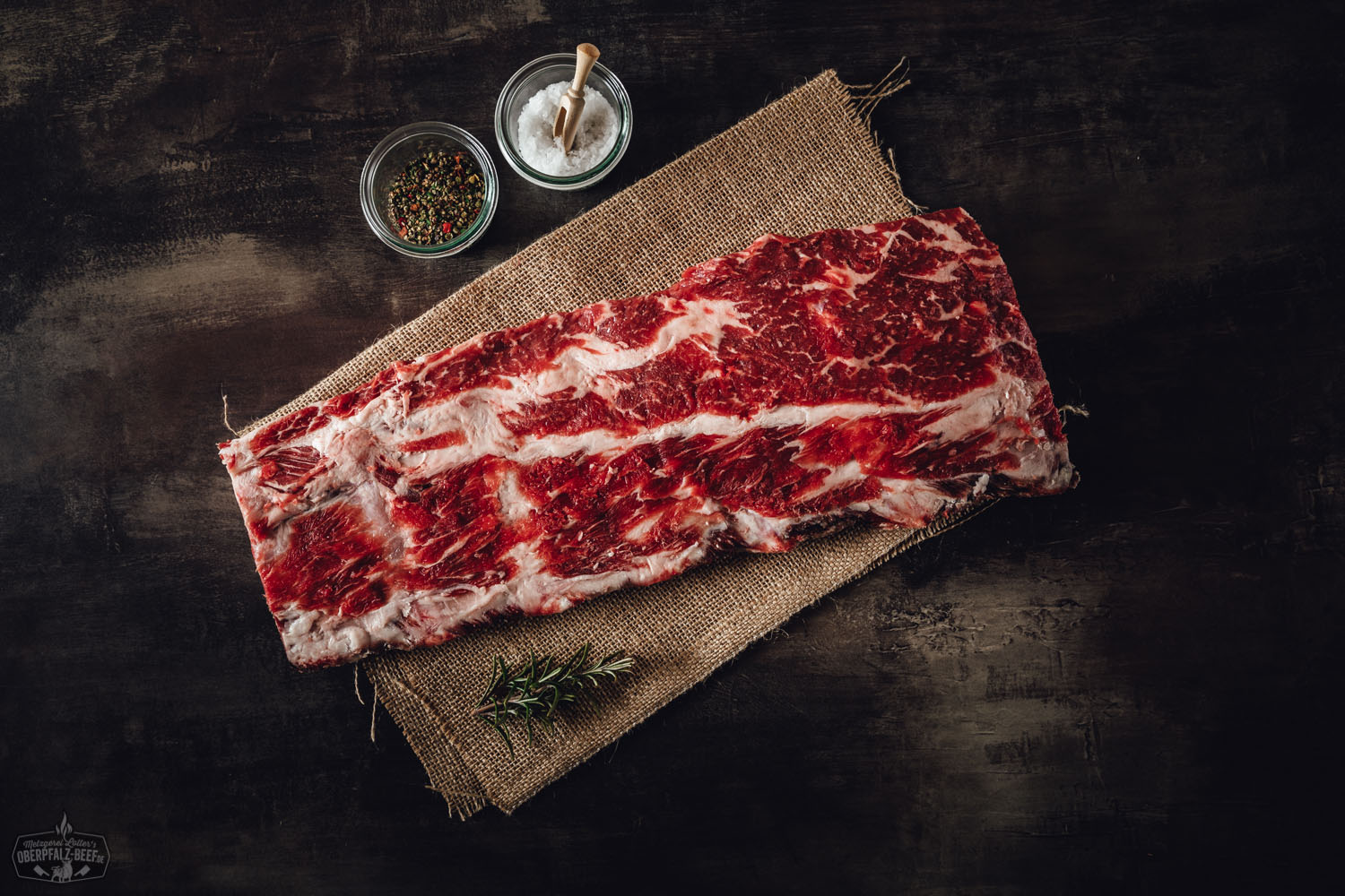 Dry Aged Back Ribs vom Oberpfalz-Rind, angerichtet auf dunklem Holzbrett mit grobem Salz, Pfefferkörnern und Rosmarinzweig – saftig gebräunte Rinderrippen mit sichtbarer Marmorierung in rustikaler Fleischpräsentation.