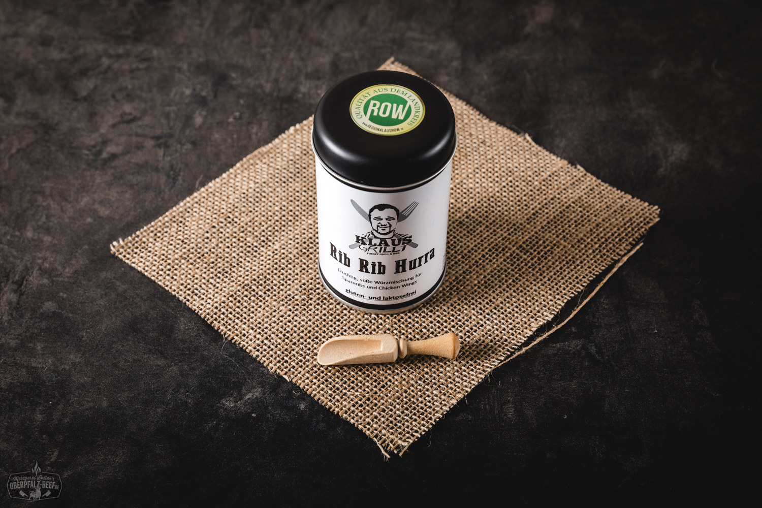 Männerhand hält Würzstreuer mit Aufschrift „Rib Rib Hurra“ über gegrilltem Fleisch auf Grillrost im Freien – BBQ-Gewürz für Rippchen beim Grillen von Oberpfalz Beef.