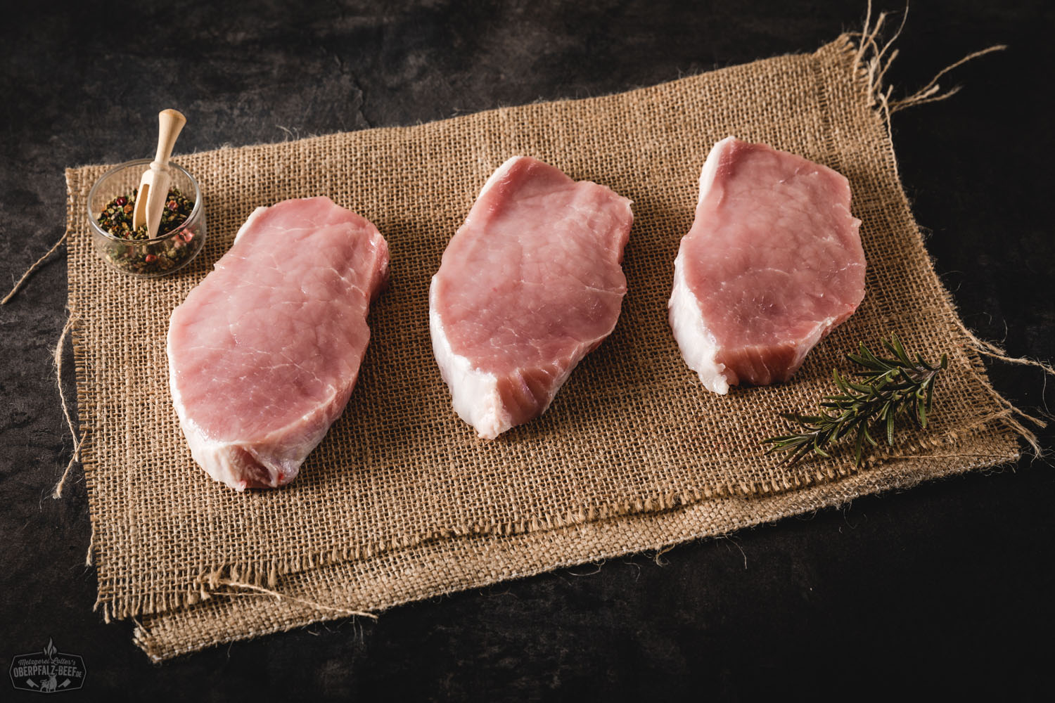 Rohe Rückensteaks vom Oberpfälzer Landschwein auf Holzbrett angerichtet, mit sichtbarer Marmorierung, frischer Rosmarinzweig und Pfefferkörner als Dekoration – hochwertiges Schweinefleisch aus regionaler Aufzucht für Grill oder Pfanne.