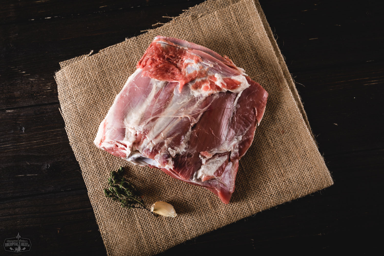 Halbe Lammschulter vom Juradistl-Lamm – regionales Weidefleisch aus der Oberpfalz Rohe halbe Lammschulter vom Juradistl-Lamm auf dunklem Untergrund, mit frischen Kräutern, Knoblauch und grobem Salz garniert – hochwertige Fleischzutat aus nachhaltiger Aufzucht für deftige Lammgerichte.