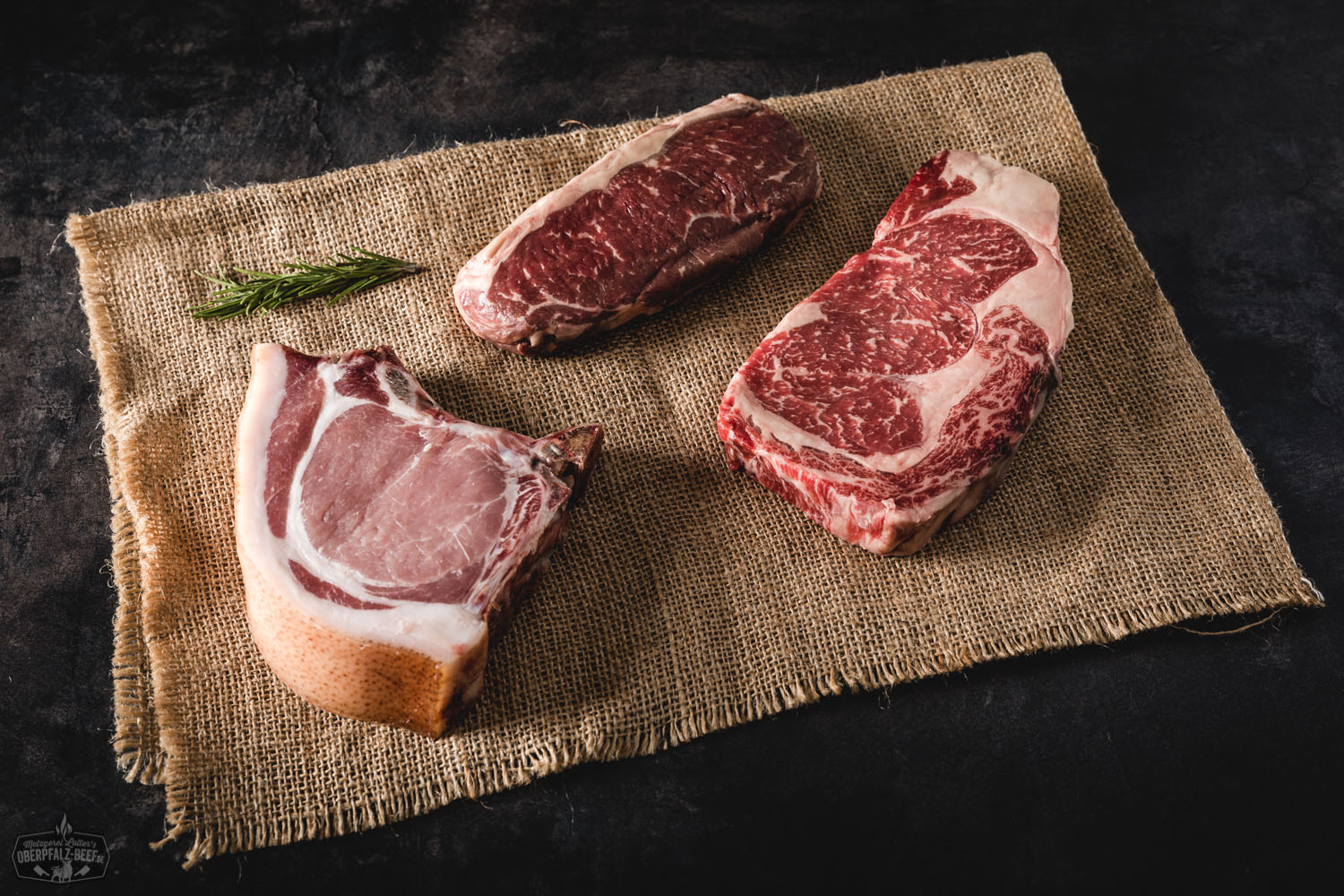 Kennenlernpaket von Oberpfalz Beef mit vakuumverpackten Rindfleisch-Stücken auf Holzuntergrund, darunter Steaks, Braten und Würstchen; beigefügt sind Produktetiketten und frische Kräuter – hochwertiges regionales Fleisch in Premium-Qualität.