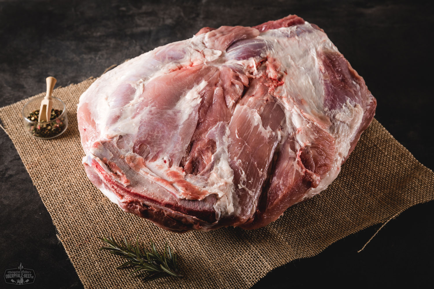 Rohe Schweineschulter mit Knochen vom Oberpfälzer Landschwein, auf einer schwarzen Schieferplatte angerichtet; das gut marmorierte Fleisch mit sichtbaren Fettstrukturen liegt neben Kräutern, Gewürzen und einem Messer – ideal für Pulled Pork Zubereitung.