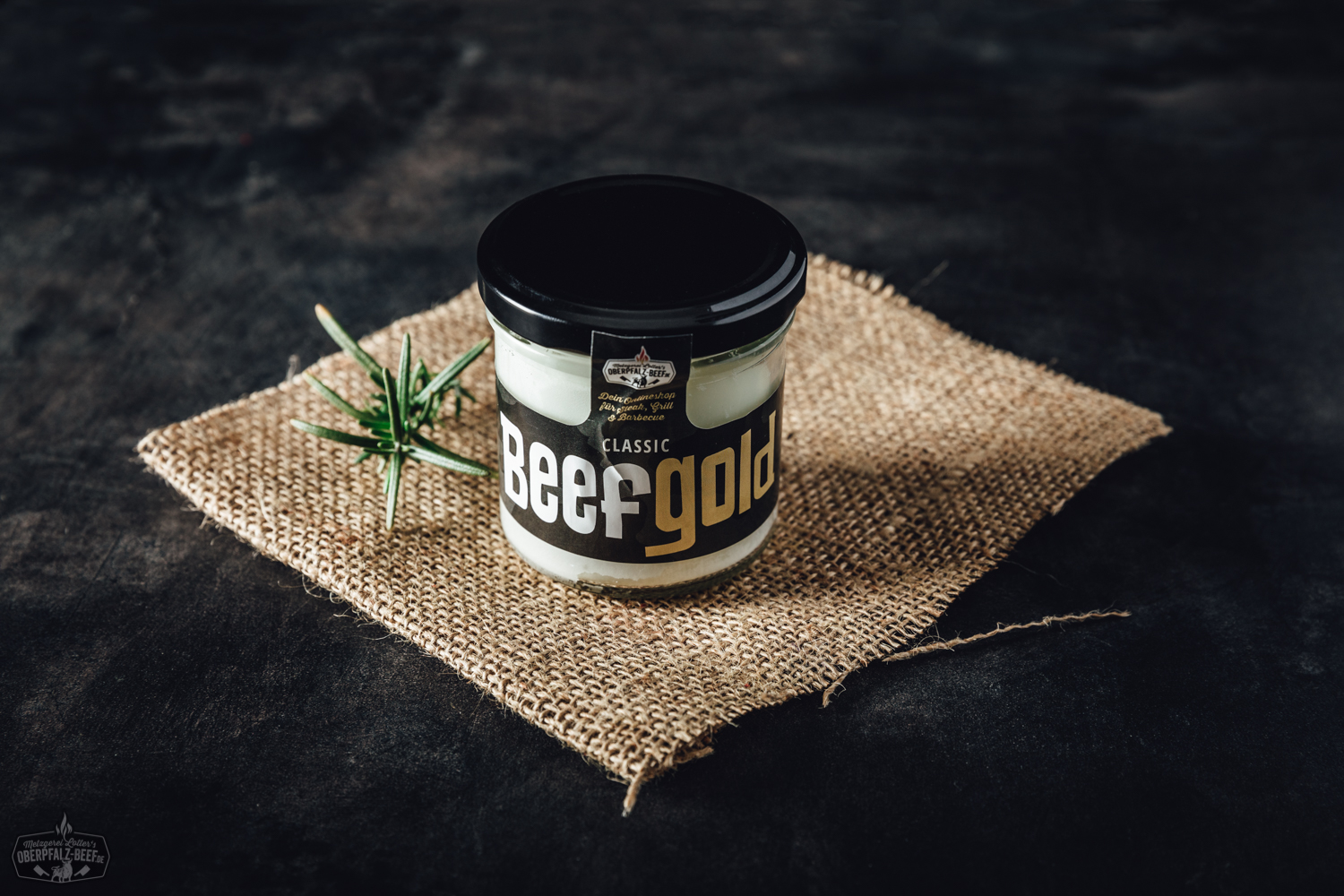 Ein geöffnetes Glas mit Beefgold Classic Rindfleisch in eigener Sauce auf hellem Untergrund, daneben ein Holzbrett mit angerichtetem Rindfleisch, frischen Kräutern und einem Messer – hochwertiges Feinkostprodukt aus der Oberpfalz für Genießer.