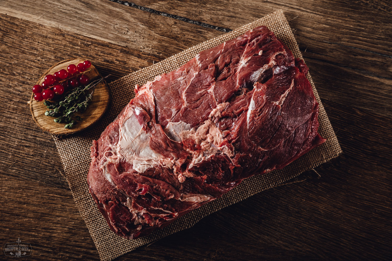 Rohes Wildschweinfleisch, zugeschnitten als Nacken ohne Knochen, auf dunklem Holzbrett angerichtet – marmoriert, frisch und saftig, ideal für Braten oder Grillgerichte; umgeben von Gewürzen wie Rosmarin, Pfeffer und Knoblauch.