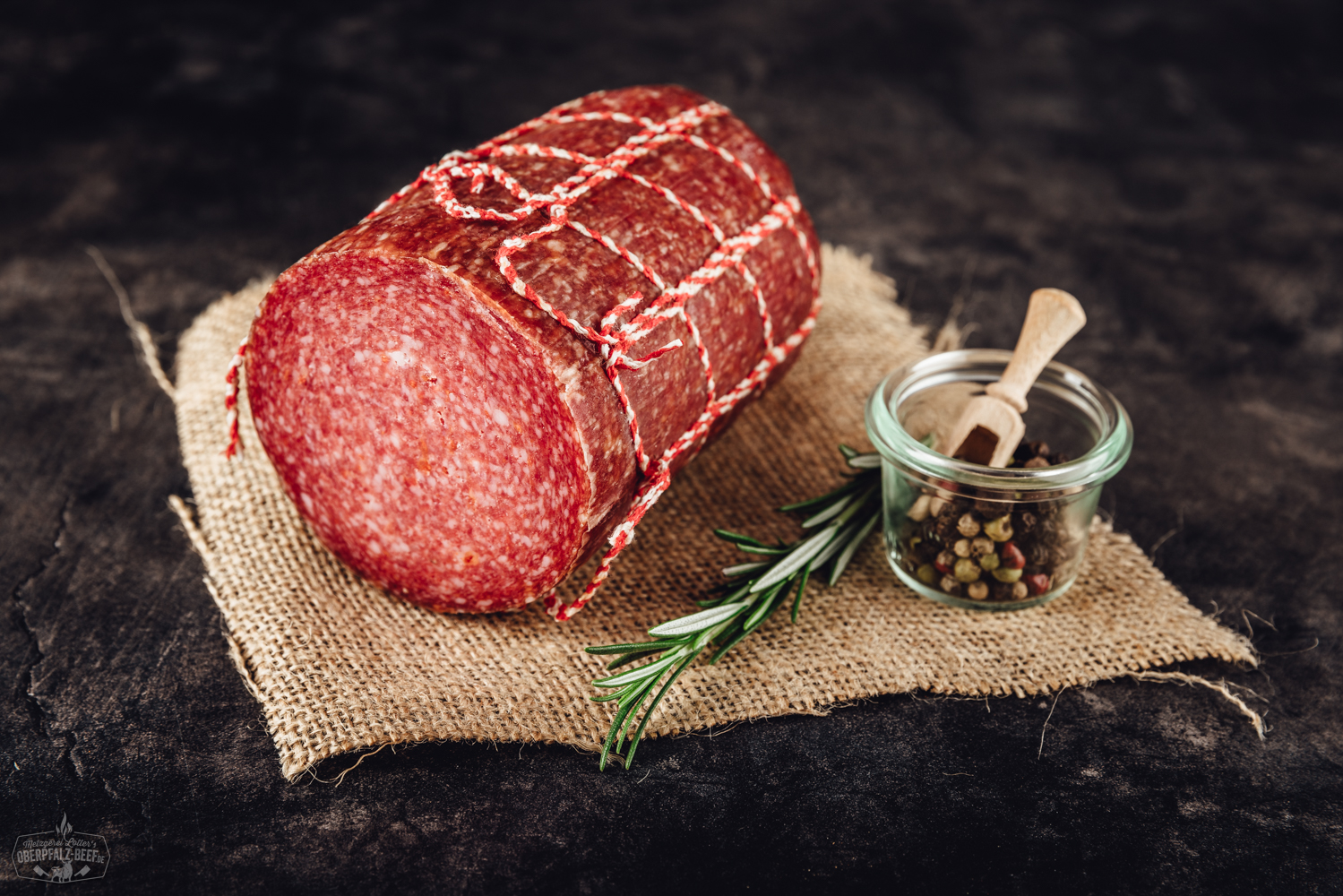 Geschnittene Scheiben von zartem, rosafarbenem Kräuterschinken auf einem Holzbrett angerichtet, dekoriert mit frischen Kräutern wie Rosmarin und Petersilie, vor hellem, neutralem Hintergrund – delikate Fleischspezialität aus der Oberpfalz.