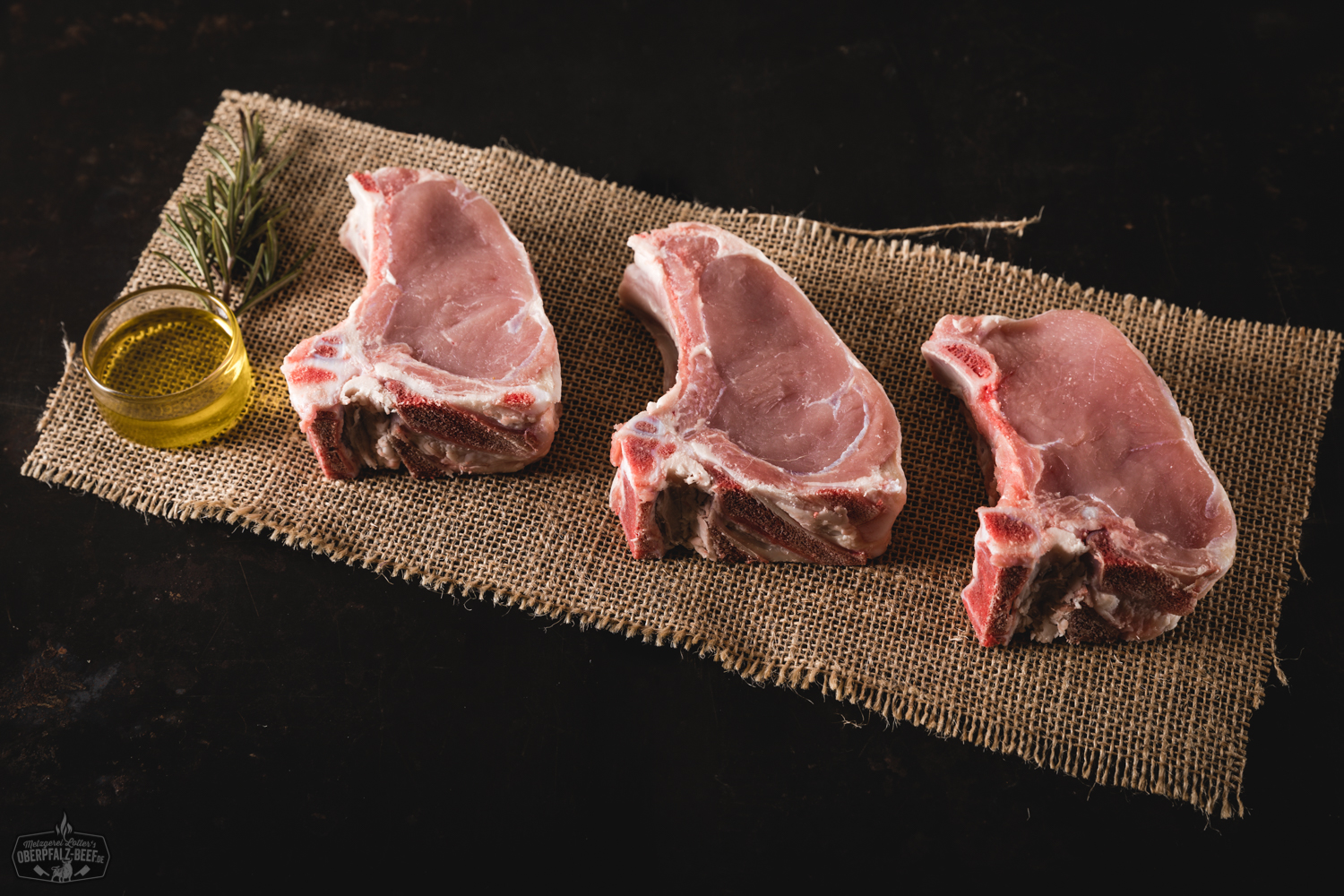 Kotelett vom Oberpfälzer Milchkalb frisch gegrillt auf Holzbrett serviert Rohes Kotelett vom Oberpfälzer Milchkalb mit hellem, zart marmoriertem Fleisch und kräftigem Knochenstück, appetitlich angerichtet auf dunkler Schieferplatte mit Gewürzen, Rosmarin und Knoblauch – hochwertige Delikatesse aus regionaler Aufzucht.