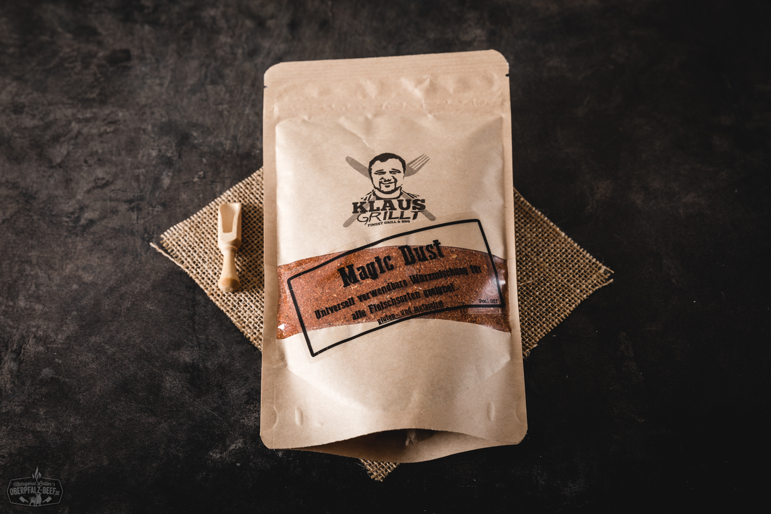 Gewürzmischung „Klaus grillt – Magic Dust“ in einem braunen Papiertütchen mit schwarzem Etikett, aufrecht stehend auf einer dunklen Holzoberfläche, im Hintergrund unscharfes Grillzubehör – aromatische BBQ-Gewürze für Fleisch, ideal fürs Grillen.