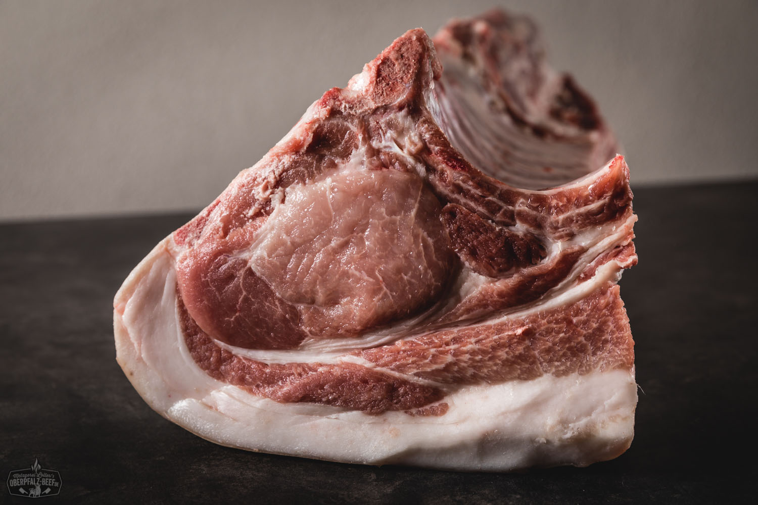 Halbes Kotelett Classic Cut vom Oberpfälzer Landschwein – Frisch aus artgerechter Haltung Frisches halbes Kotelett vom Oberpfälzer Landschwein im Classic Cut, roh auf schwarzem Untergrund präsentiert, mit sichtbarer Marmorierung und Fettrand – hochwertiges Schweinefleisch für Grill oder Pfanne.