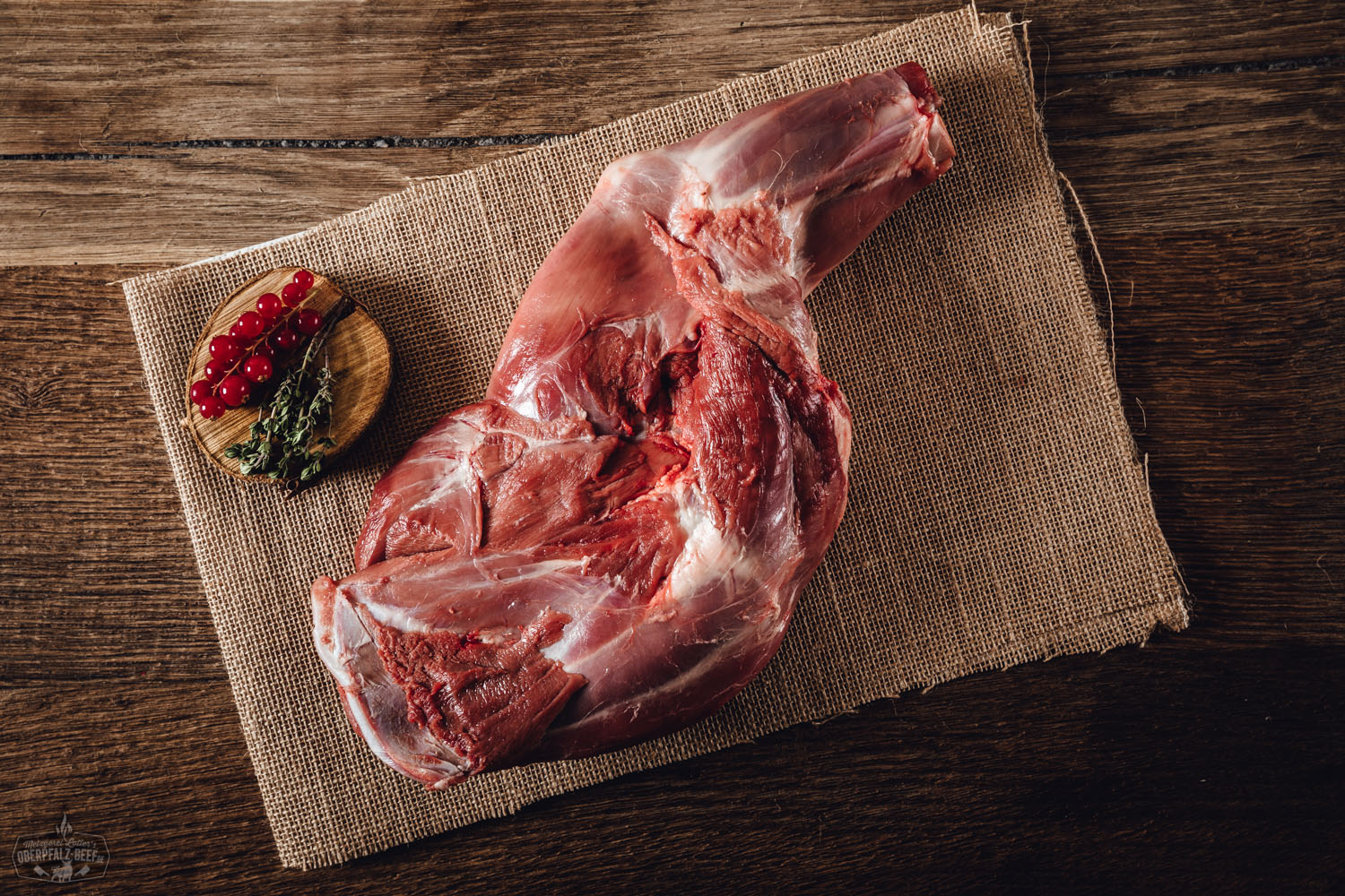 Schulter mit Knochen vom Oberpfälzer Reh – Wildfleisch in Premiumqualität Roher Rehschulterbraten mit Knochen vom Oberpfälzer Reh, auf einem dunklen Schneidebrett angerichtet, umgeben von frischen Kräutern, Wacholderbeeren, Knoblauch und Rosmarinzweigen – hochwertiges Wildfleisch, ideal für Braten oder Schmorgerichte.