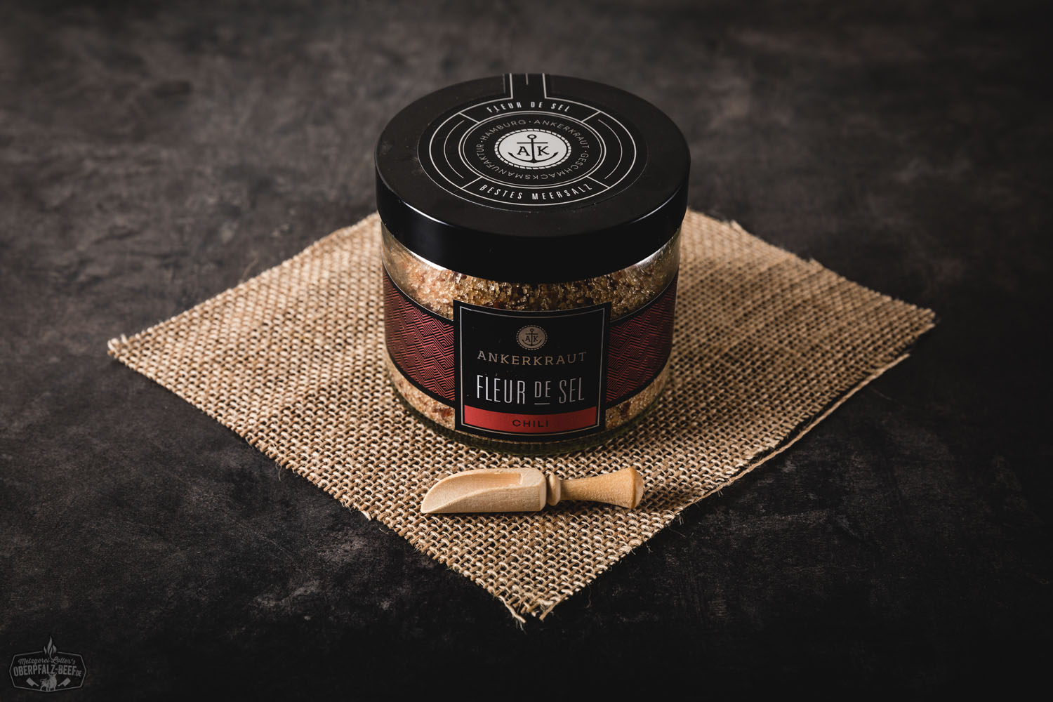 Glas mit Ankerkraut Fleur de Sel Chili Gewürzmischung vor dunklem Hintergrund, daneben grobes Meersalz mit Chiliflocken und getrockneten Chilischoten auf schwarzem Untergrund – hochwertige Gewürzzubereitung für Fleisch, Grillgerichte und feurige Speisen.