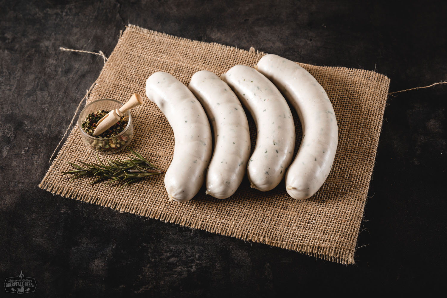 Frische Weißwürste mit Petersilie und Senf auf traditionellem Holzteller Mehrere traditionelle bayerische Weißwürste liegen angerichtet auf einem weißen Teller, begleitet von einer Brezel, süßem Senf und frischer Petersilie, im Hintergrund ein weiß-blauer Tischläufer – typisch bayerisches Frühstück.