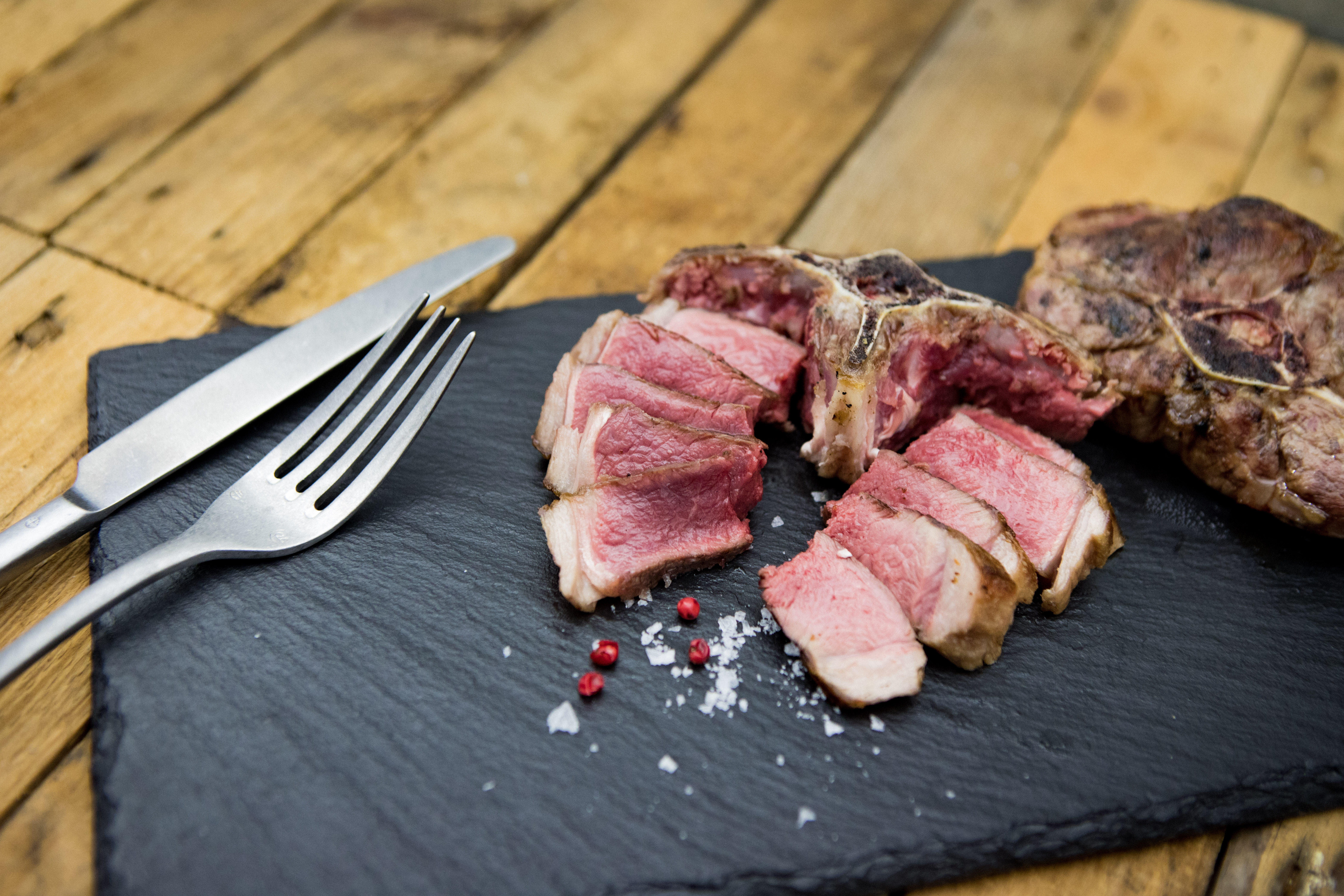 Zarte Lammkoteletts aus dem Hals vom Juradistl-Lamm – regionales Premiumfleisch aus der Oberpfalz Flasche Spyridoulas 100% natives Olivenöl extra auf hellem Holzuntergrund, daneben frische grüne Oliven und ein kleiner Olivenzweig – authentisches griechisches Olivenöl für hochwertige mediterrane Küche.