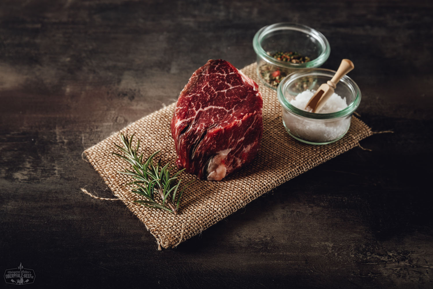 Dry Aged Filetsteak von der alten Kuh, roh und sorgfältig pariert auf dunkler Steinplatte angerichtet, umgeben von grobem Meersalz, grünem Rosmarinzweig und schwarzem Pfeffer – hochwertiges Rindfleisch in Premium-Qualität für Gourmet-Grillgerichte.