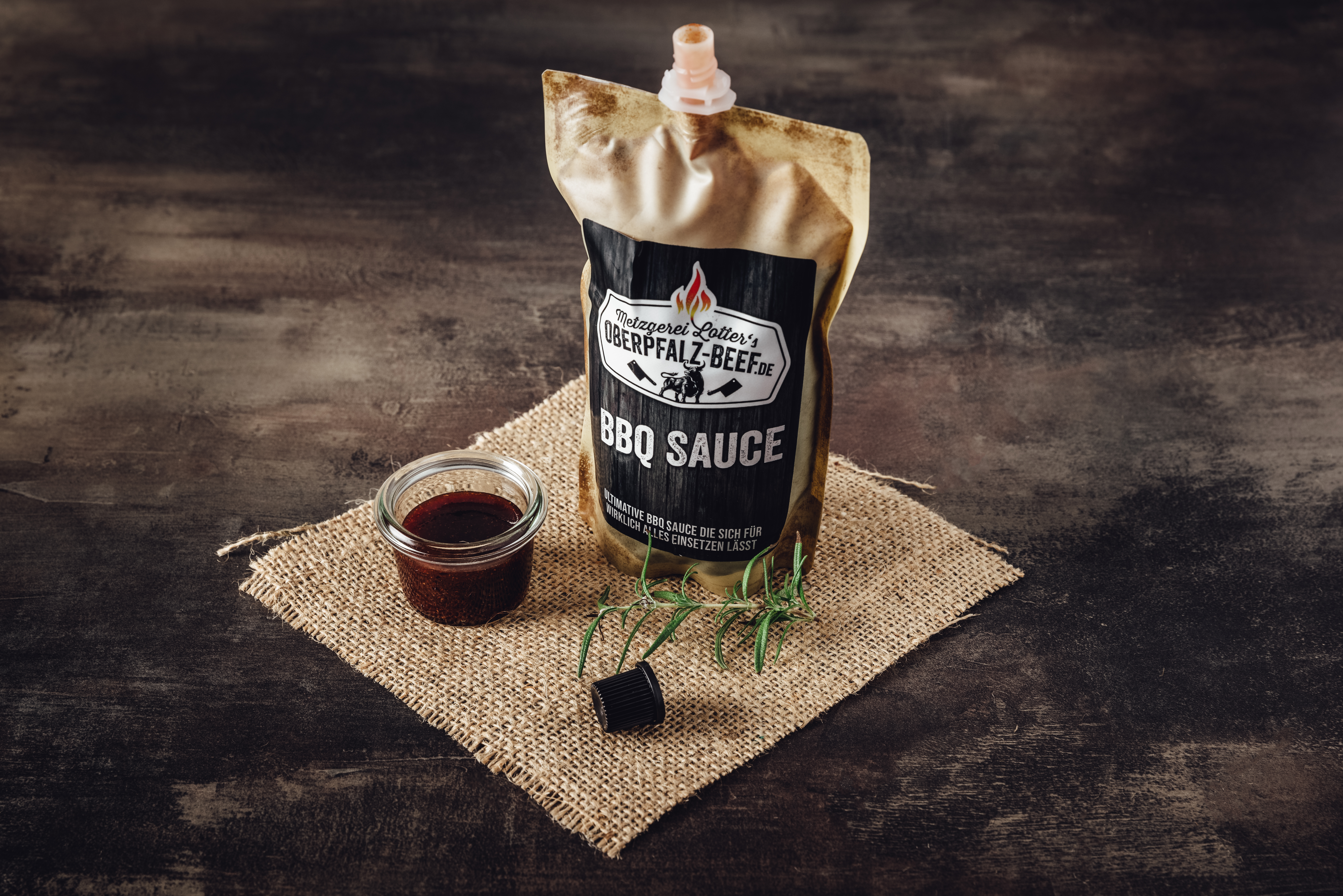 BBQ Sauce in Glasflasche – Hochwertige Zutaten und intensiver Geschmack Flasche mit BBQ Sauce auf Holztisch, mit rotem Etikett und Zutatenlisten im Hintergrund, ideal für Grillabende und Fleischgerichte.