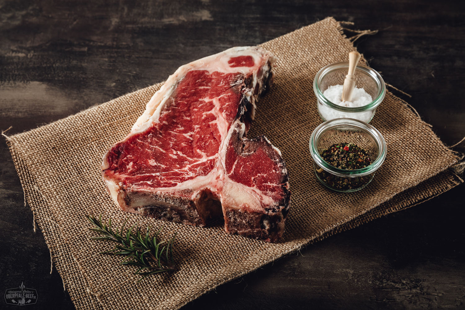 Auf einem Holzbrett liegt ein Stück zart marmoriertes Kalbfleisch, die Nuss vom Oberpfälzer Milchkalb, roh und frisch zugeschnitten; daneben frische Rosmarinzweige, Pfefferkörner und Meersalz – hochwertige Zutaten für Feinschmecker und Fleischliebhaber.