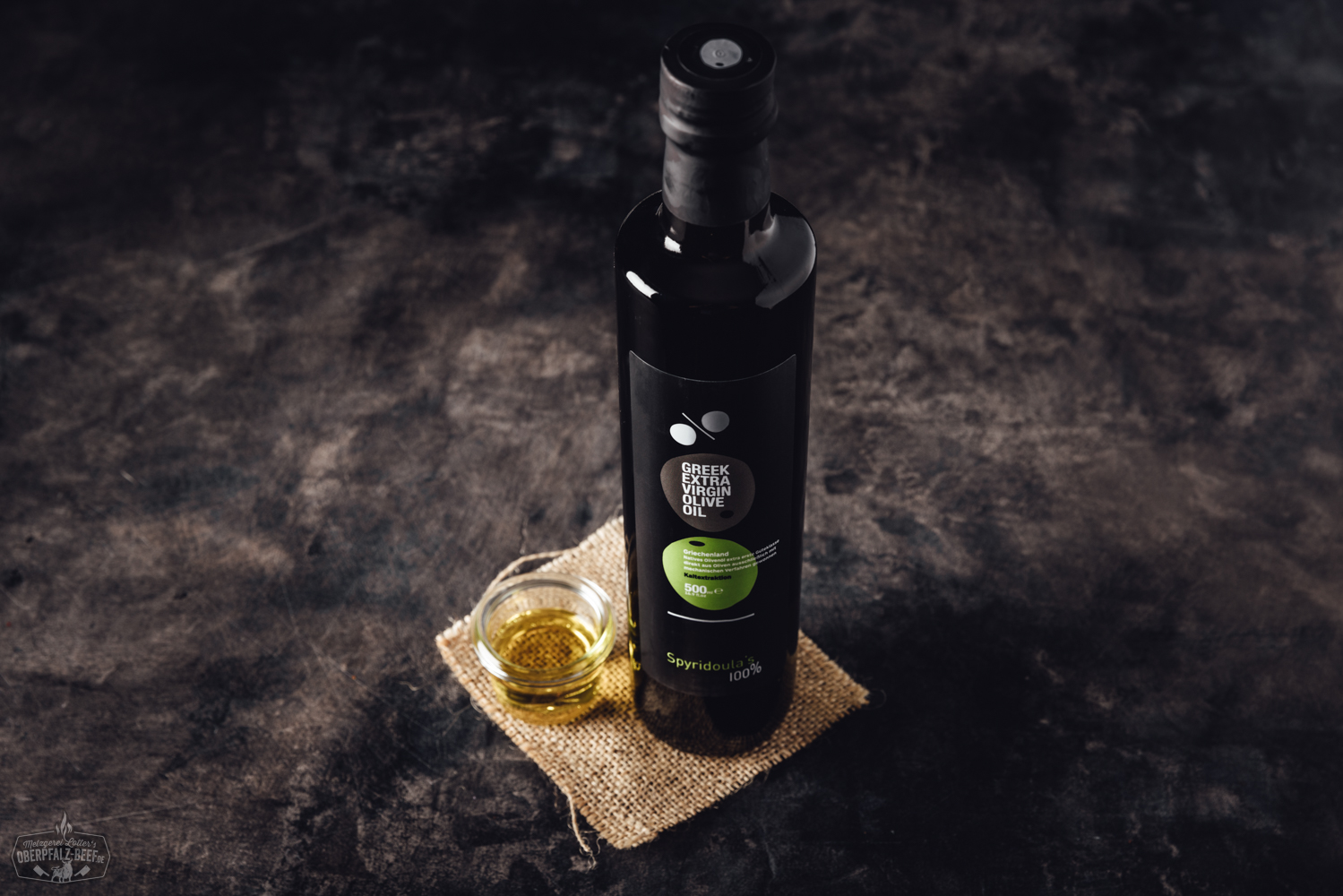 Spyridoulas natives Olivenöl extra – Hochwertiges griechisches Olivenöl in eleganter Flasche Steakmesser aus der Dick 1905 Serie im Oberpfalz-Beef-Stil mit edlem Holzgriff, liegend auf einem rustikalen Holzbrett vor dunklem Hintergrund – hochwertiges Fleischmesser für präzises Schneiden von Steak und Fleisch.