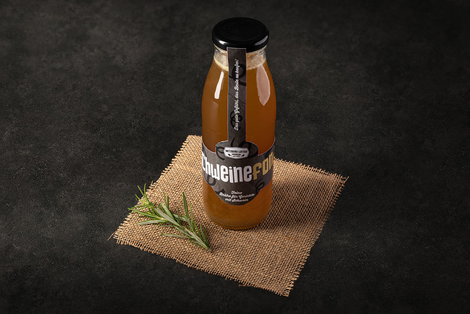 Ein Einmachglas mit Schweinefond, gefüllt mit klarer, goldbrauner Brühe, steht auf einem dunklen Untergrund; daneben frische Zutaten wie Sellerie, Karotte, Lauch und Gewürze – rustikale Präsentation für hochwertiges Fleisch- und Suppenprodukt.