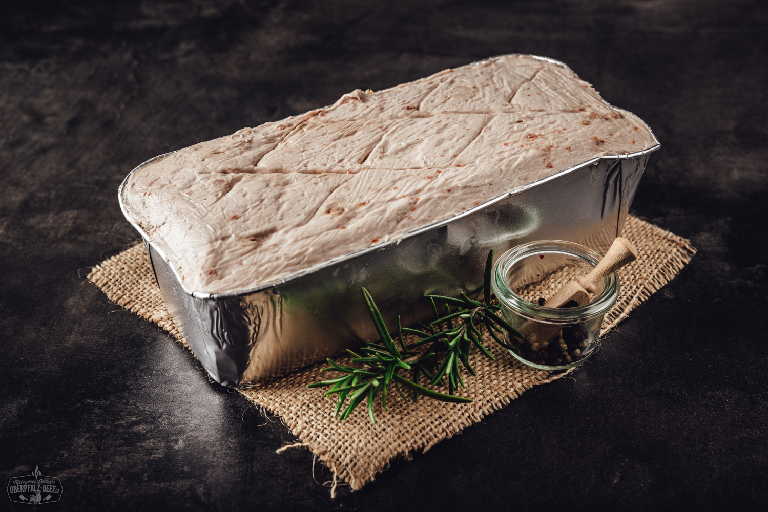 Nahaufnahme eines vakuumverpackten Blocks Pizzakäse zum Selber-Aufbacken von Oberpfalz Beef auf dunklem Hintergrund; deutlich sichtbare, fein geriebene Käsemischung in transparenter Kunststoffverpackung mit gelbem Etikett und schwarzer Schrift.