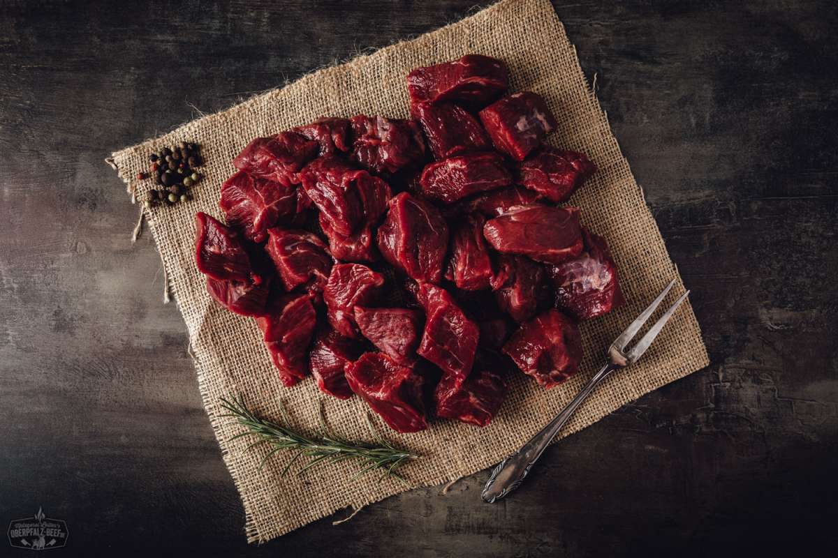 Handgeschnittenes Rindergulasch vom Oberpfalz-Rind – Frisch & Regional Handgeschnittenes Rindergulasch vom Oberpfalz-Rind, roh und in gleichmäßige Stücke geschnitten, auf einem dunklen Holzbrett angerichtet mit frischen Rosmarinzweigen und grobem Meersalz, ideal für hochwertiges Gulasch oder Schmorgerichte.