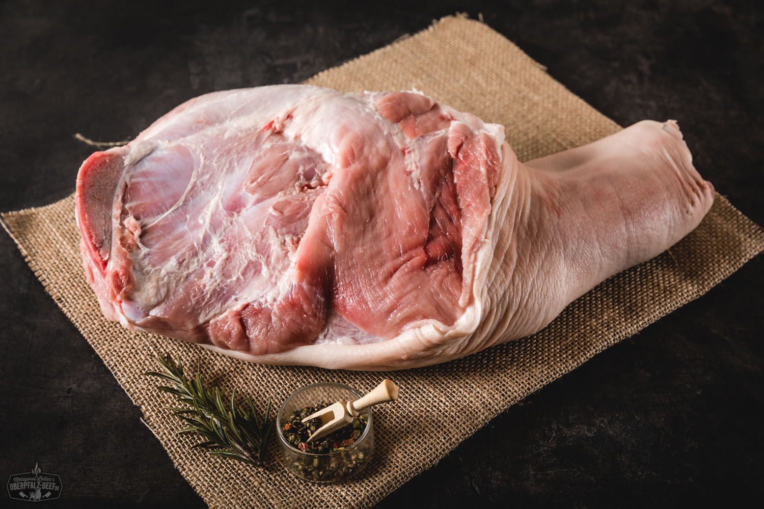 Rohe Spanferkelschulter mit Knochen vom Oberpfälzer Landschwein auf dunkler Unterlage, frisch zugeschnitten mit sichtbarer Marmorierung, eignet sich ideal zum Braten oder Grillen.