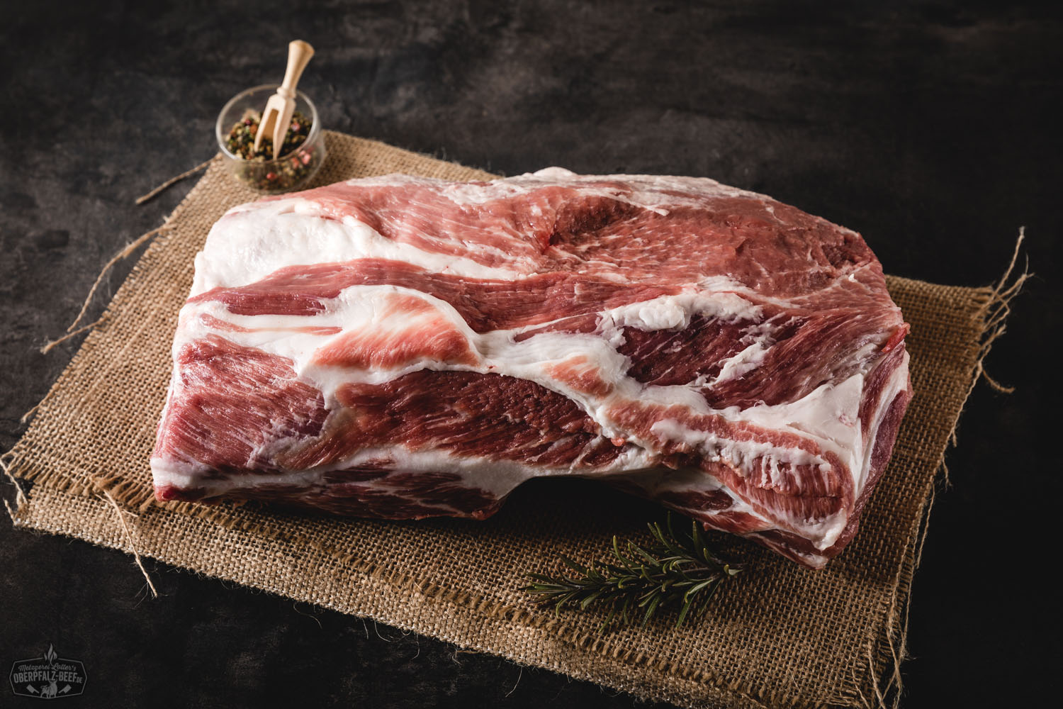 Roher Schweinehals ohne Knochen auf schwarzem Untergrund, marmoriert mit feinen Fettadern, ideal vorbereitet für Pulled Pork; daneben Gewürze und frische Kräuter zur Zubereitung – hochwertige Nahaufnahme für BBQ und Fleischliebhaber.