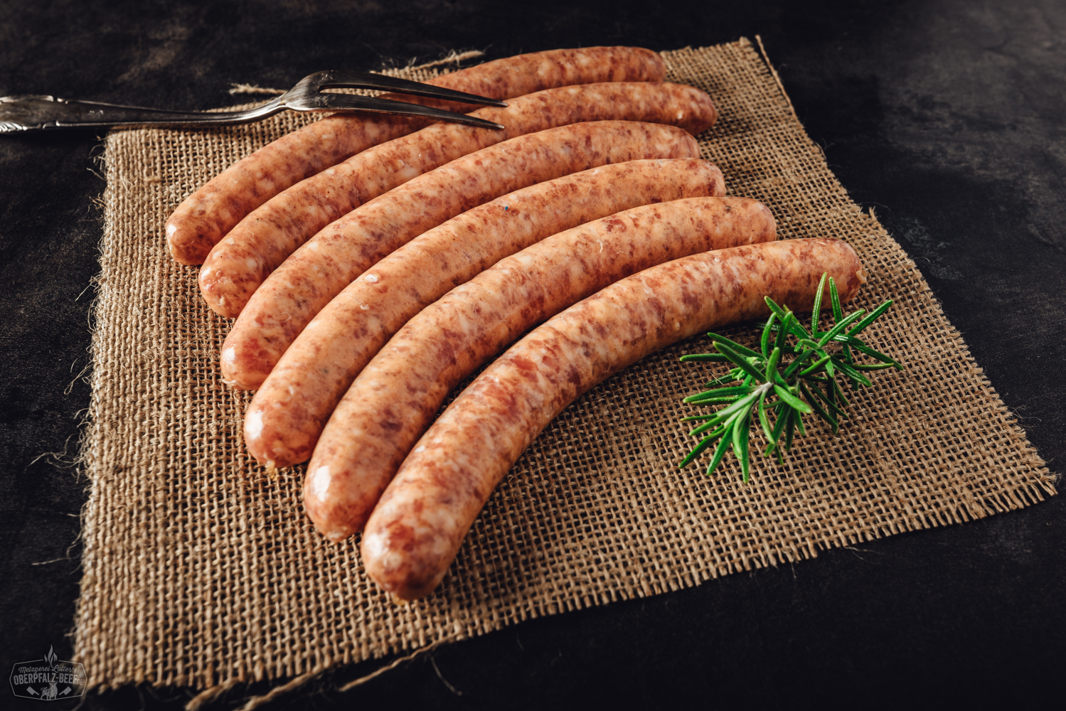 Vier grobe Wildbratwürste mit sichtbaren Kräutern und Gewürzen liegen nebeneinander auf einer dunklen Schieferplatte, begleitet von Rosmarinzweigen – hochwertige, rustikale Wildwurst aus der Oberpfalz, ideal für Grillfans und Wildfleischliebhaber.