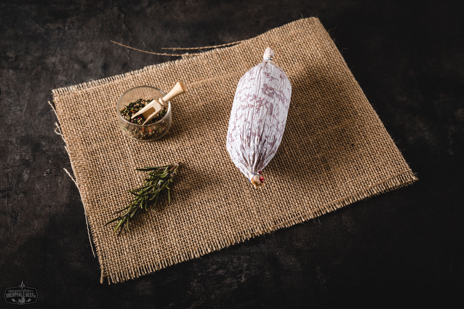 Hausgemachte Rindersalami – luftgetrocknet & regional aus der Oberpfalz Hausgemachte Salami am Stück auf dunklem Holzbrett mit angeschnittenem Ende, das die grobe, fleischige Textur zeigt; daneben frische Kräuter und Gewürze, stimmungsvolles Licht betont die rustikale, hochwertige Fleischqualität.
