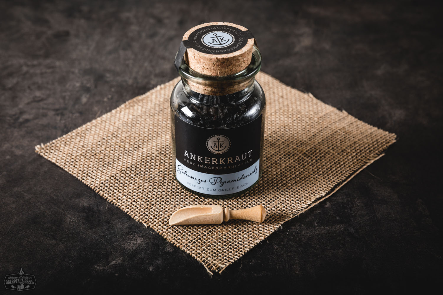 Glasgewürzdose mit schwarzem Pyramidensalz von Ankerkraut, auf einem dunklen Untergrund arrangiert, grobe Salzflocken sichtbar vor der Dose platziert – hochwertige Gewürzpräsentation für exklusives Kocherlebnis.