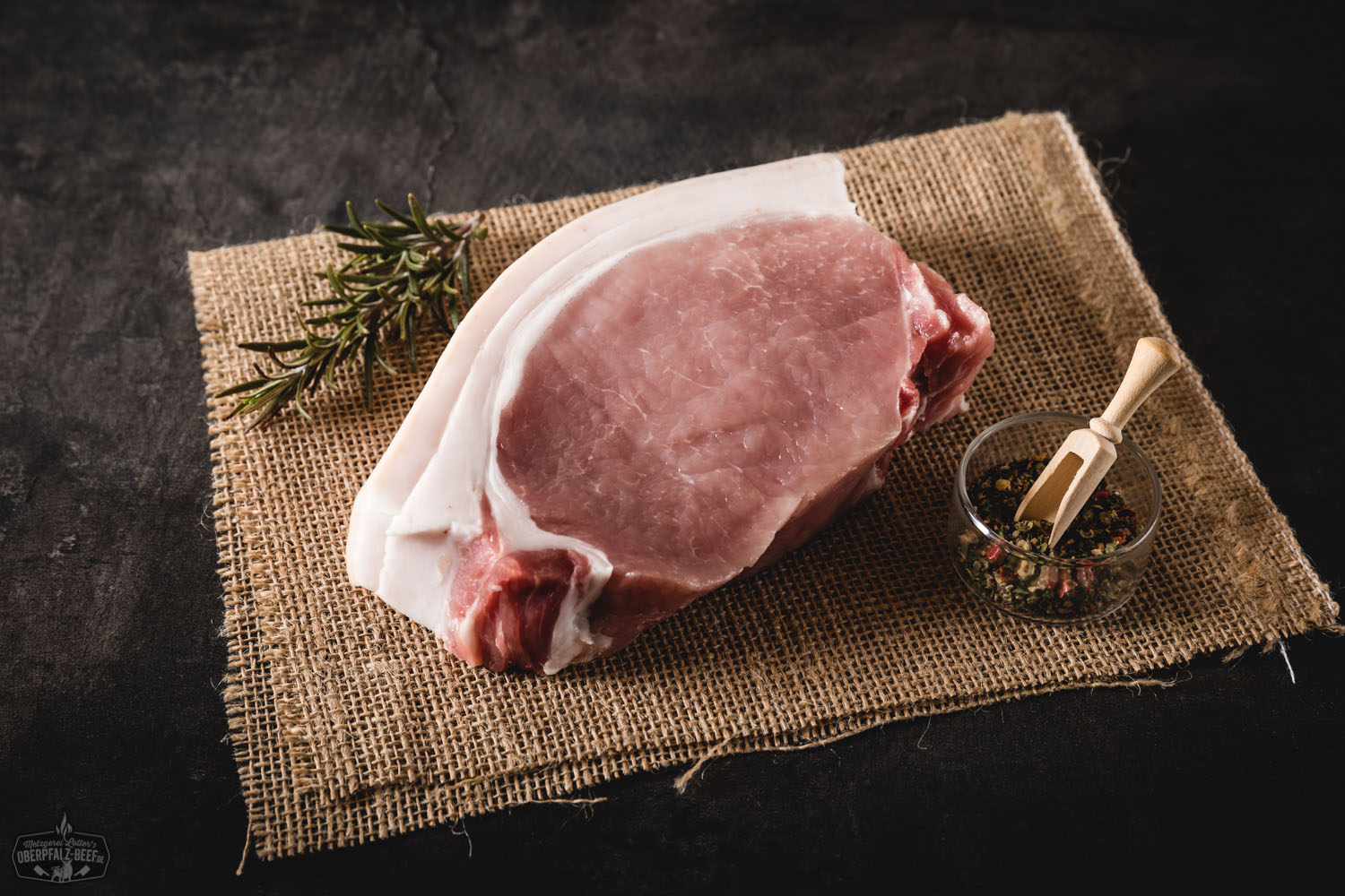 Frisches Kotelettsteak vom Oberpfälzer Landschwein roh auf dunklem Untergrund, gut marmoriert mit sichtbarem Knochen, appetitlich angerichtet mit Kräutern und Gewürzen – hochwertiges Schweinefleisch für Grill oder Pfanne.