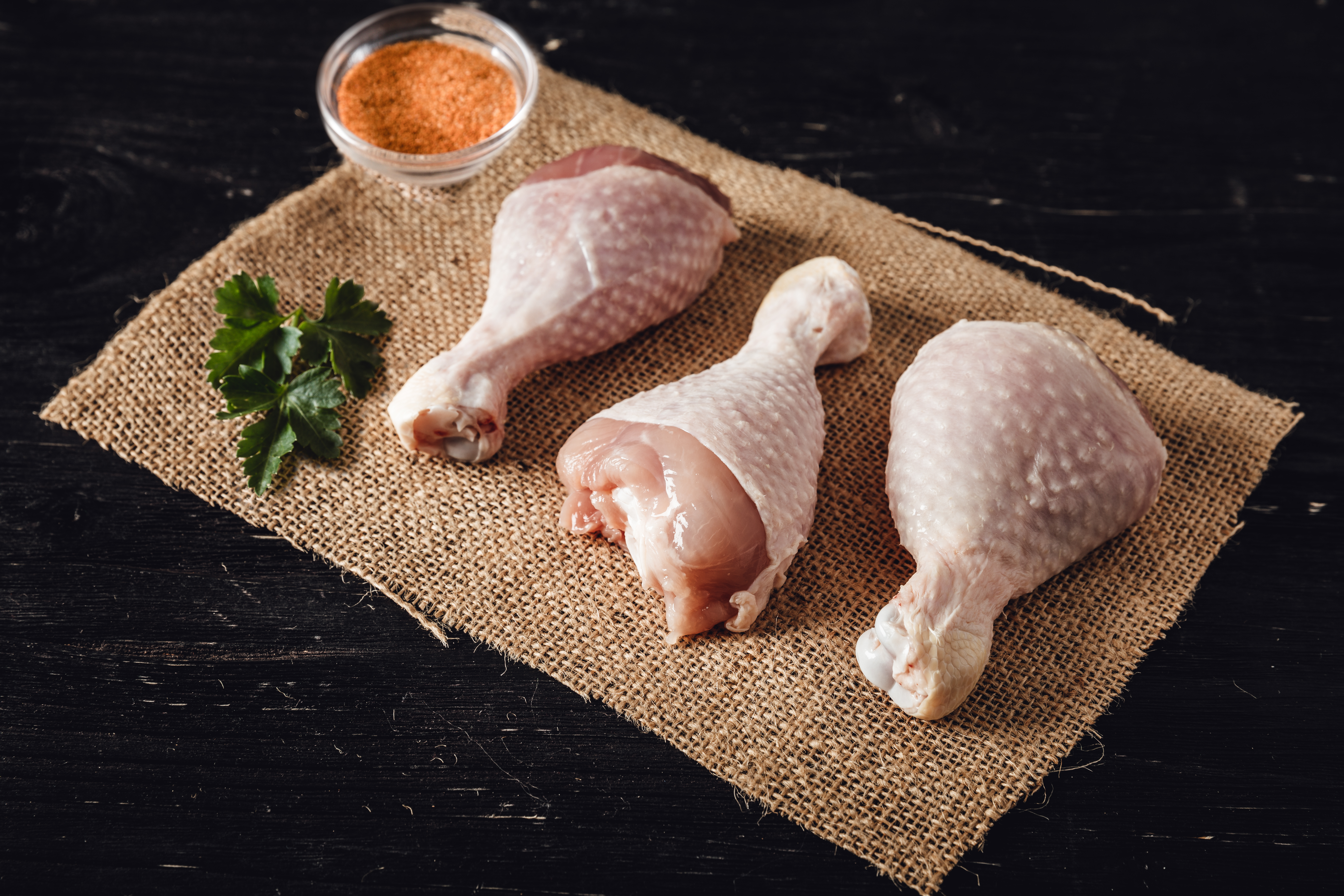 Rohe Hähnchen-Drumsticks vom Oberpfälzer Bauernhähnchen liegen auf einem dunklen Brett, umgeben von frischen Kräutern, Gemüse und Gewürzen; hochwertiges, regionales Geflügelfleisch für gesundes, nachhaltiges Kochen.