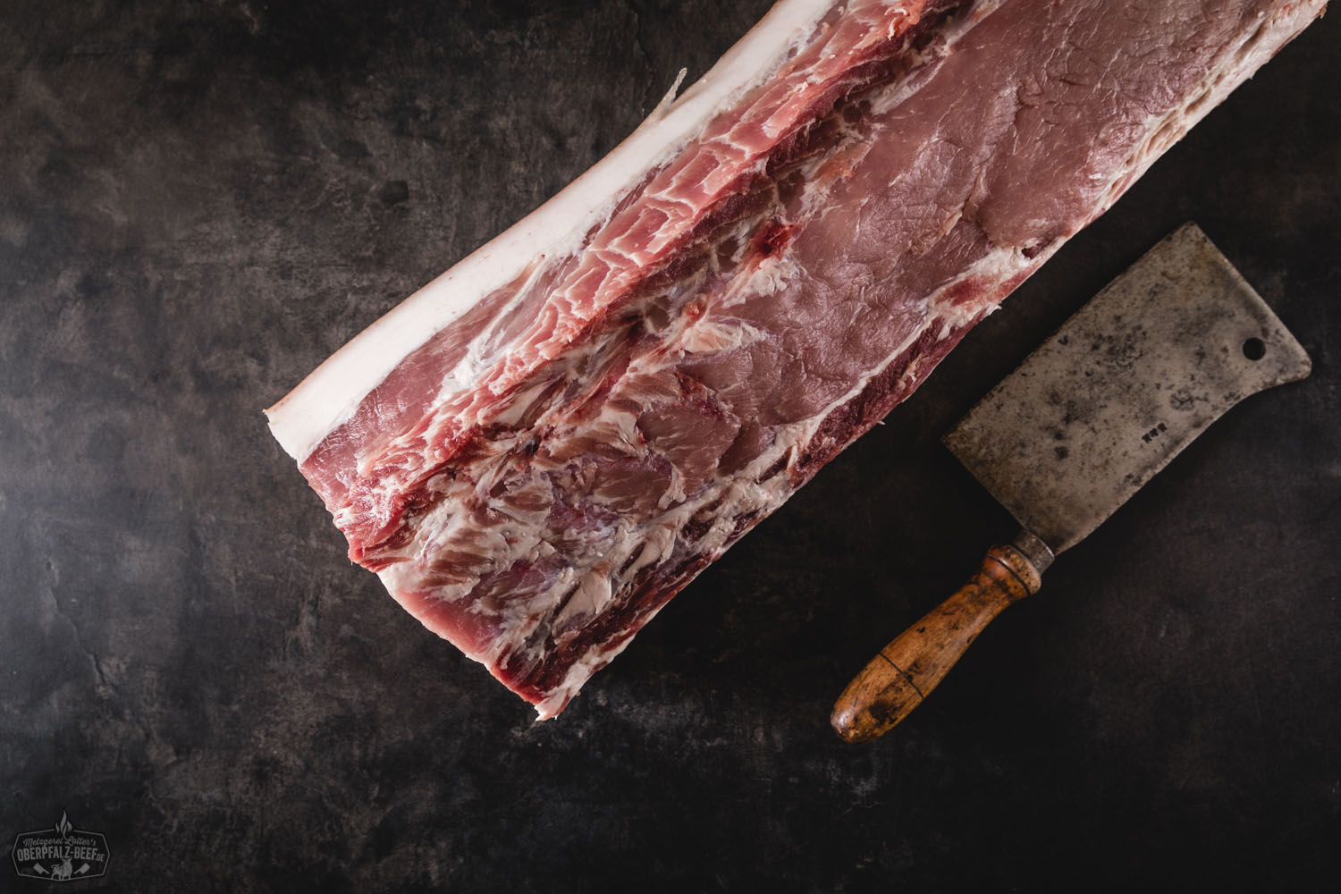 Dry Aged Kotelett mit Schwarte vom Oberpfälzer Landschwein – hochwertiges Premiumfleisch Ganzes Dry Aged Kotelett mit Schwarte vom Oberpfälzer Landschwein, roh und frisch auf dunklem Untergrund präsentiert; gut marmoriertes Fleisch mit Knochen, typisch für hochwertige Schweinefleisch-Cuts aus nachhaltiger Regionalzucht.