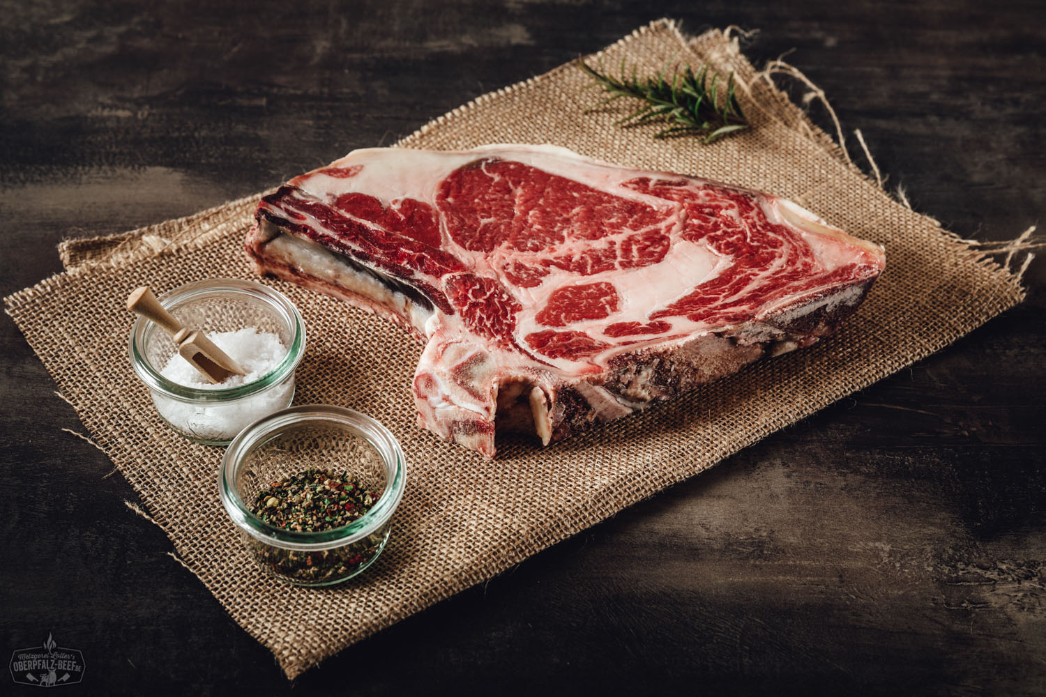 Dry Aged Entrecôte Rib-Eye am Knochen auf dunklem Untergrund; das marmorierte Rindfleisch weist eine kräftige rote Farbe und feine Fettäderchen auf, ideal zur Zubereitung als Steak – hochwertiges Premiumfleisch für echte Fleischliebhaber.