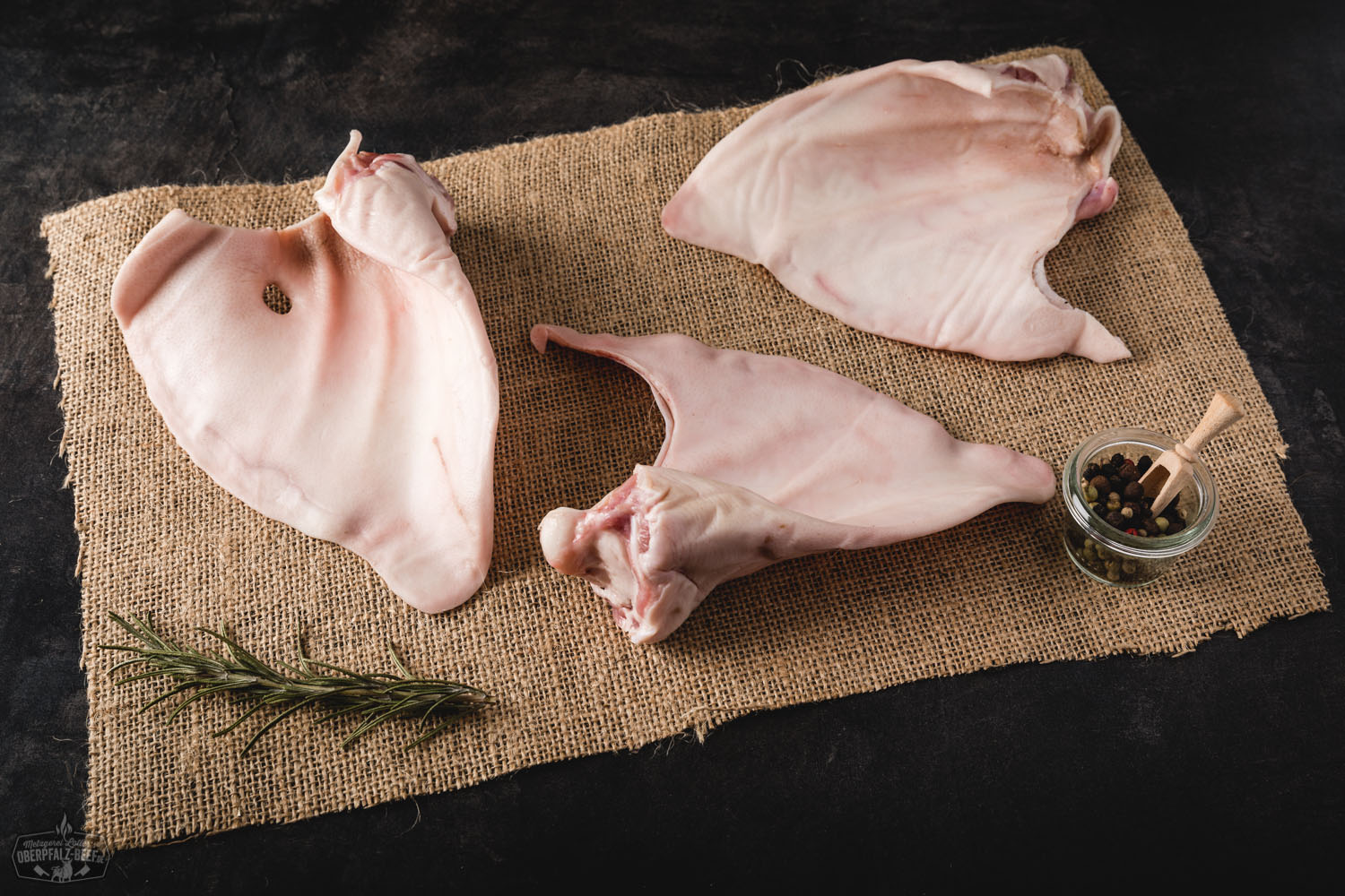 Ohren vom Oberpfälzer Landschwein – Regionales Schweinefleisch direkt vom Erzeuger Roh geschnittene Schweineohren vom Oberpfälzer Landschwein liegen appetitlich auf einem dunklen Holzbrett, umgeben von grobem Salz, Pfeffer und frischen Kräutern – hochwertige regionale Fleischspezialität aus Bayern.