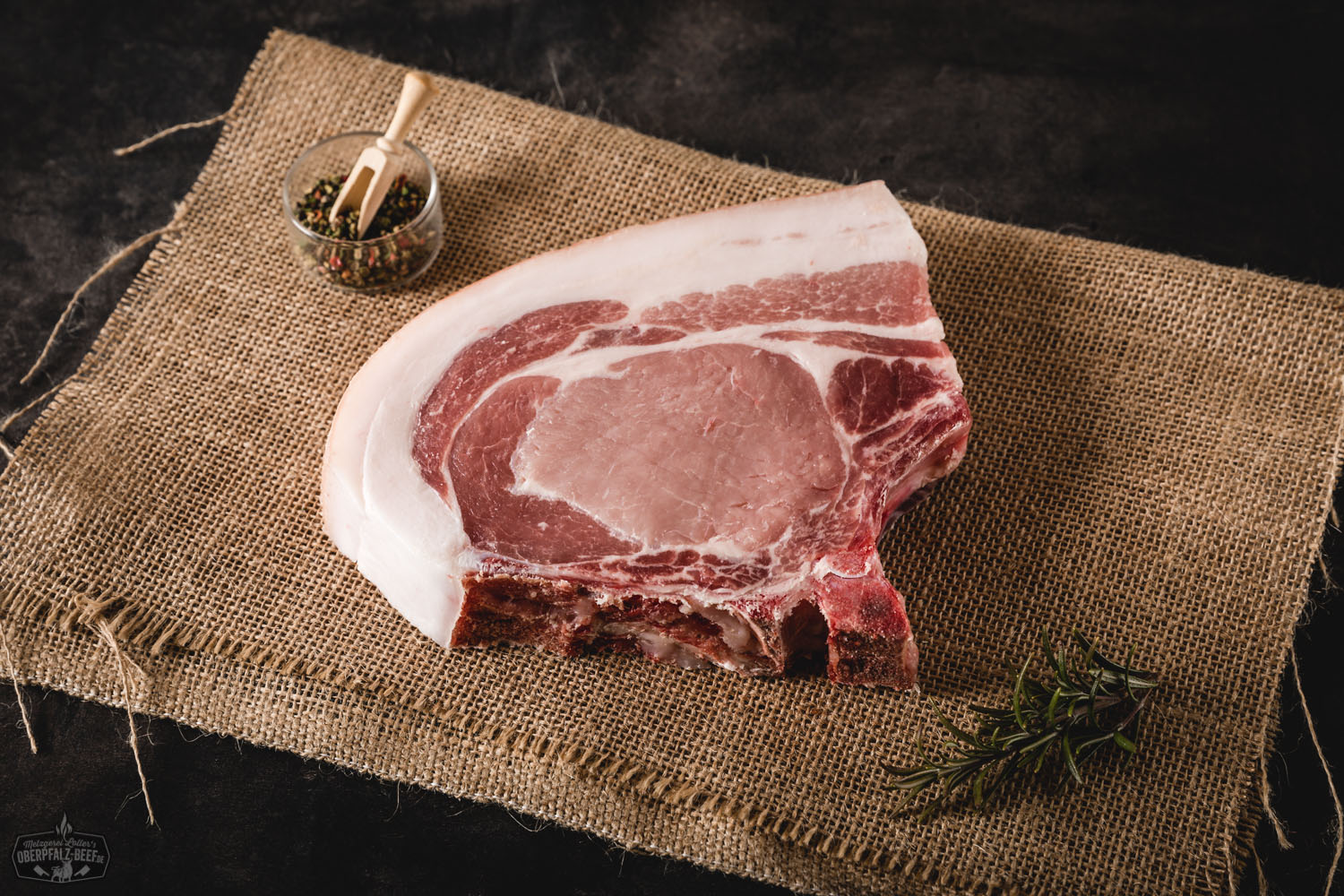Dry Aged Kotelett-Steak vom Oberpfälzer Landschwein – Premium Schweinefleisch Dry Aged Kotelett-Steak vom Oberpfälzer Landschwein auf dunklem Untergrund, roh präsentiert mit feiner Marmorierung, sichtbarem Knochen und frischem Thymian garniert – hochwertiges Premiumfleisch für Grill oder Pfanne.