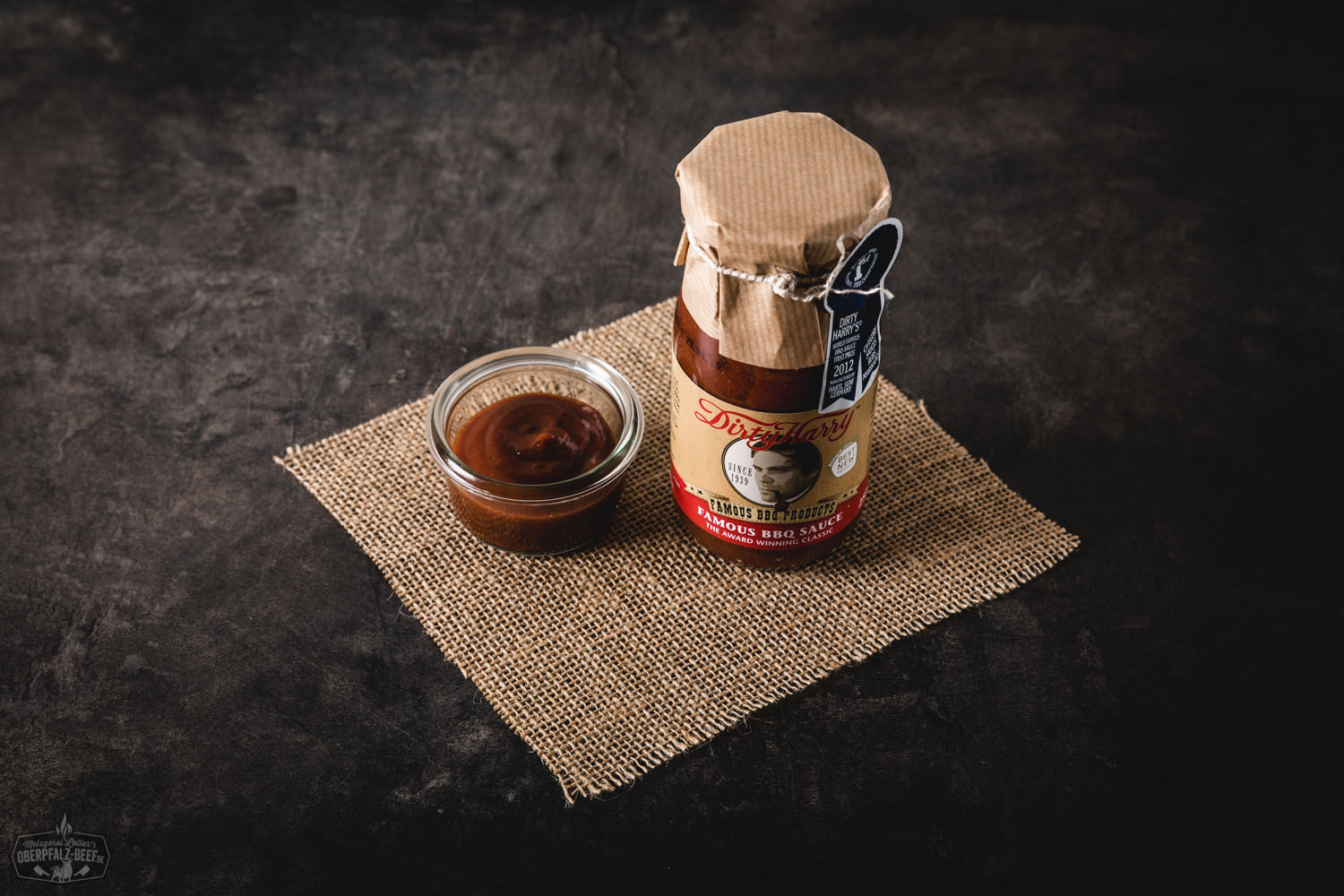 Dirty Harry Famous BBQ Sauce – Würzige Barbecue-Sauce im Glas mit Kultfaktor Flasche „Dirty Harry Famous BBQ Sauce“ mit schwarzem Etikett, roter Schrift und Western-Motiv; Produkt steht auf rustikalem Holztisch mit Grillzubehör im Hintergrund – ideal für BBQ, Grillen und Fleischgerichte.