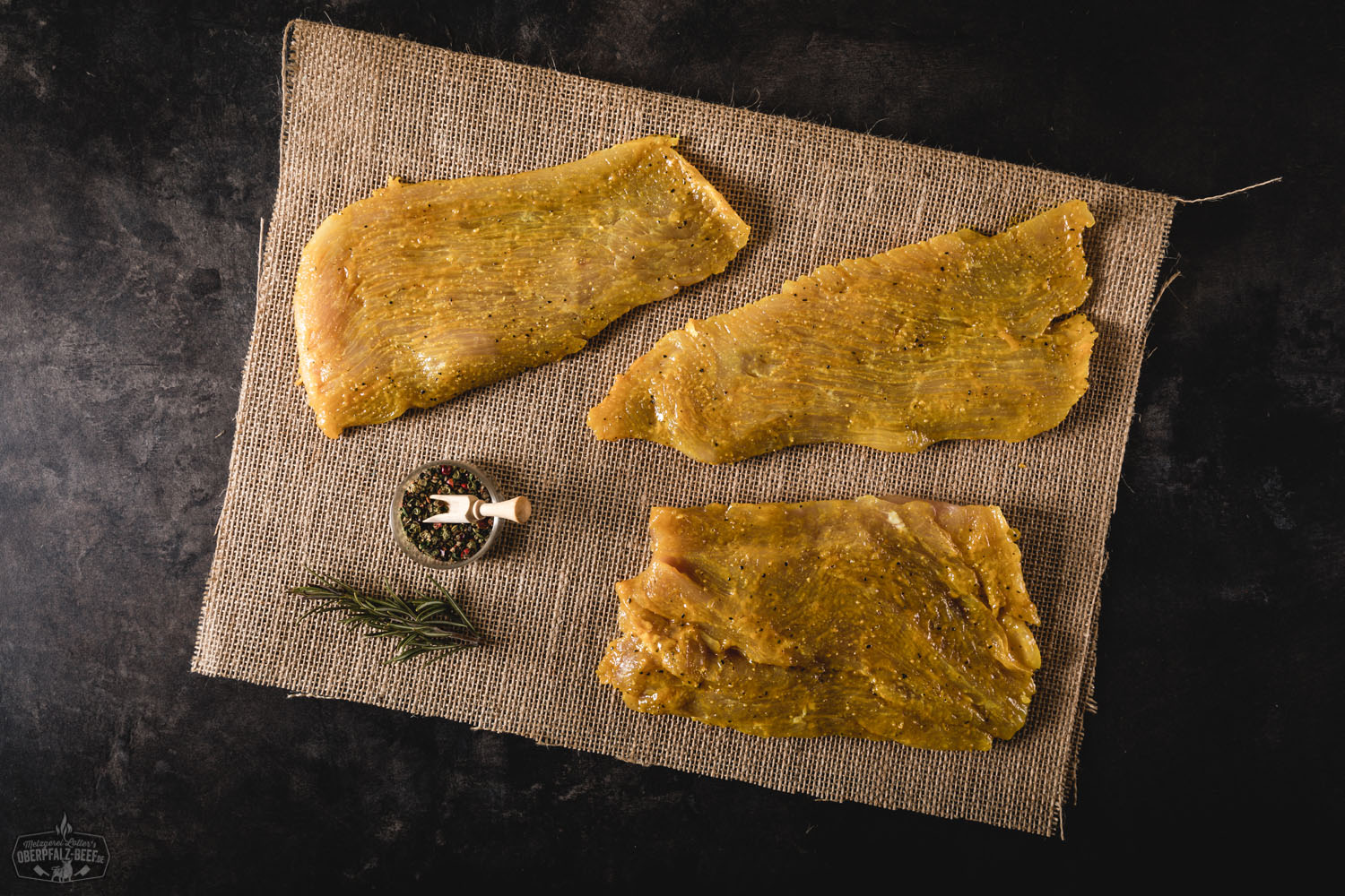 Drei gewürzte Putensteaks roh auf einem dunklen Holzbrett angerichtet, dekoriert mit grobem Pfeffer, Rosmarinzweigen, Chili, Salzflocken und Knoblauch – hochwertiges Frischfleisch für Grill oder Pfanne.