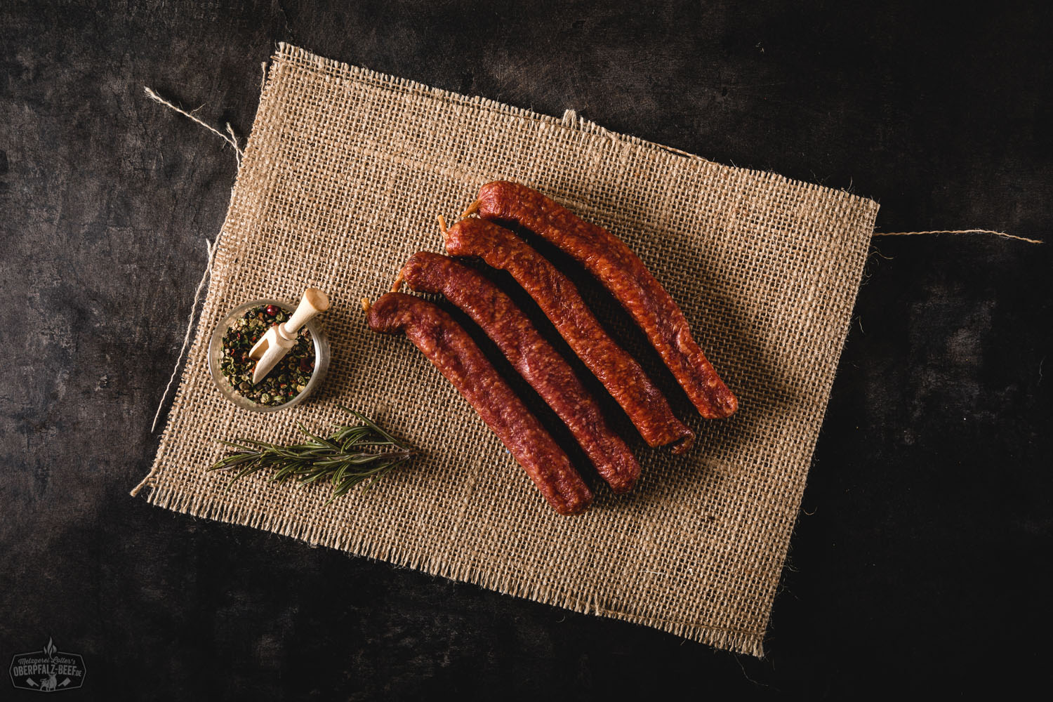Zwei traditionelle, dunkelrote Landjäger-Würste aus Rindfleisch hängen an einer Schnur vor rustikalem Holzhintergrund – Delikatess-Wurst aus der Oberpfalz, luftgetrocknet und herzhaft im Geschmack.