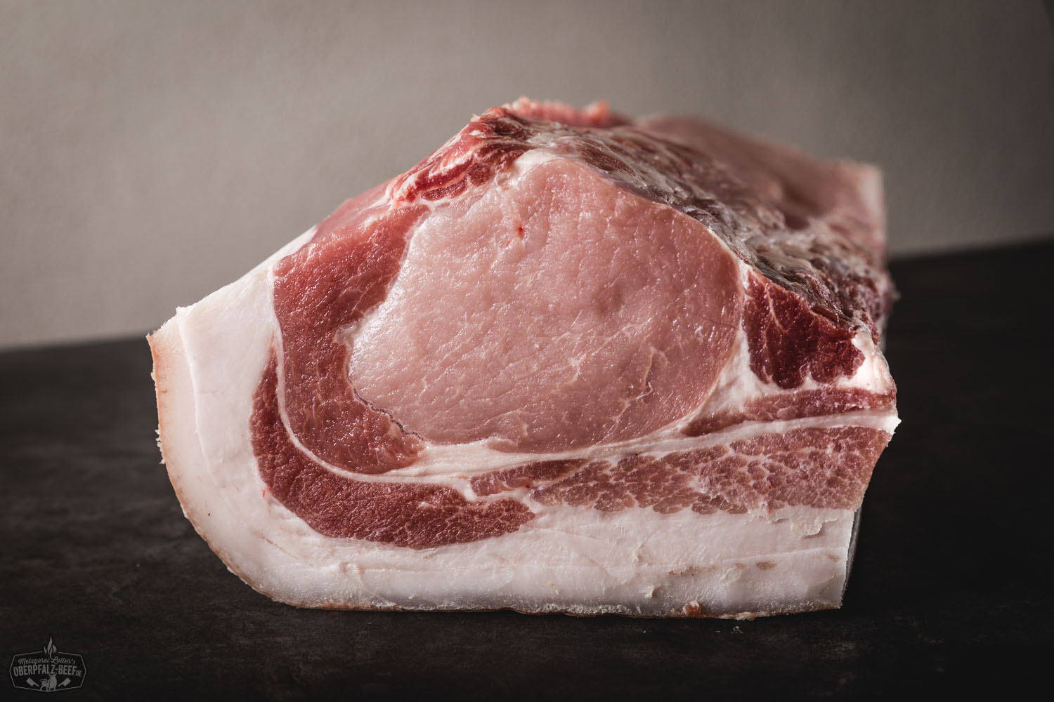 Dry Aged Kotelett mit Schwarte vom Oberpfälzer Landschwein – regionales Premiumfleisch Ganzes Dry-Aged Kotelett mit Schwarte vom Oberpfälzer Landschwein, roh auf dunklem Untergrund präsentiert, marmoriertes Fleisch mit ausgeprägtem Fettrand – hochwertiges Stück Fleisch für Grill oder Ofen.