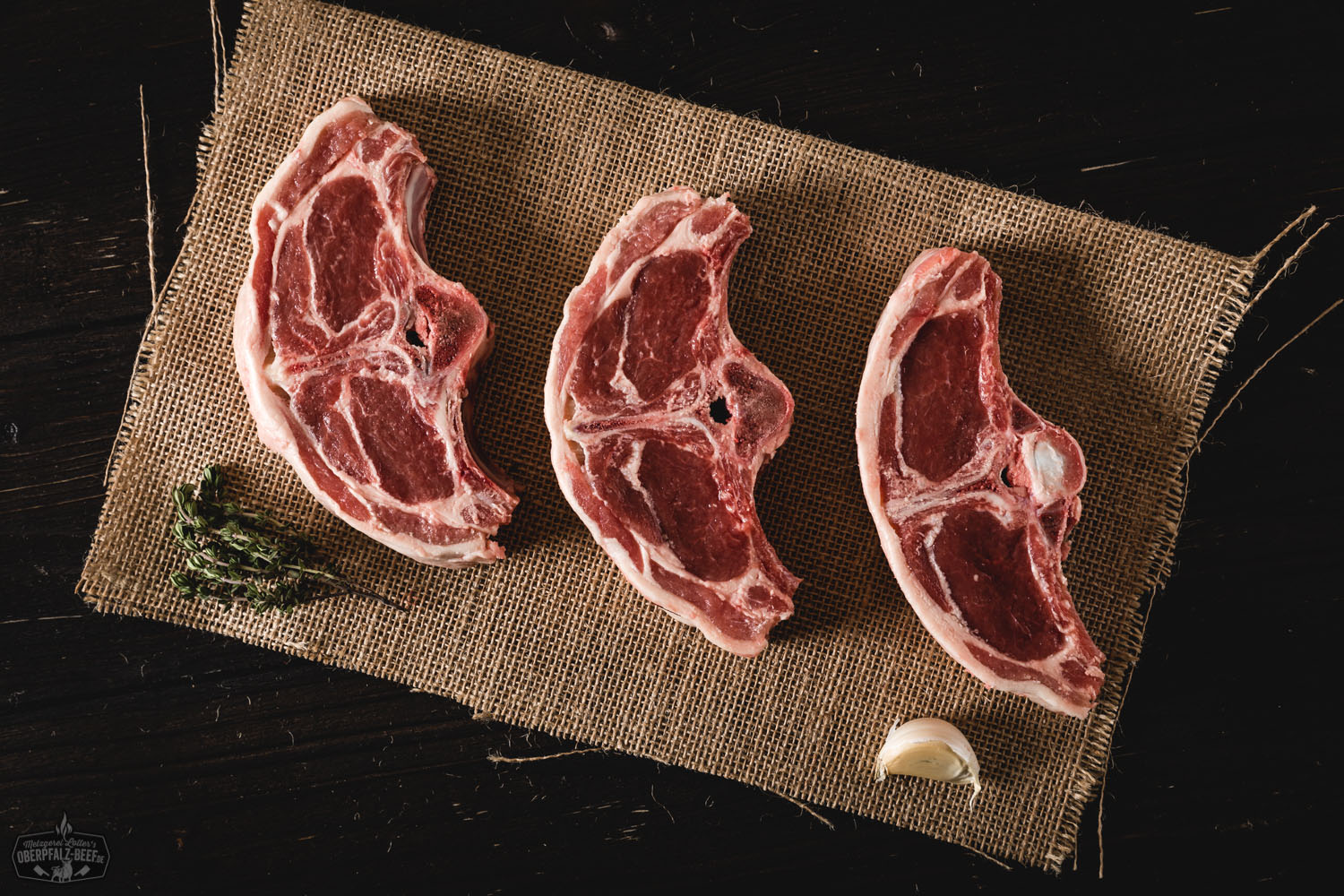 Lammkoteletts aus dem Rücken, roh und mit Knochen, liegen auf dunklem Untergrund und sind von frischen Kräutern und grobem Salz umgeben – hochwertiges Lammfleisch für Grill oder Pfanne.