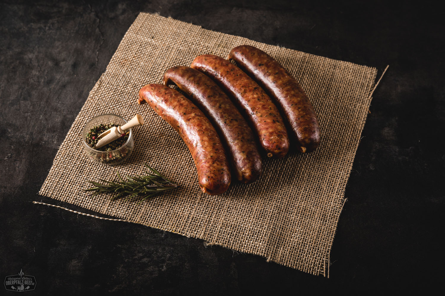 Bauernseufzer Wurst frisch gegrillt auf rustikalem Holzbrett mit Senf und Brot Vier grob gekörnte, gebräunte Bauernseufzer-Würste aus Schweinefleisch liegen nebeneinander auf einem Holzbrett, umgeben von frischen Kräutern, Zwiebelringen und buntem Pfeffer – rustikal angerichtet für eine herzhafte Brotzeit.