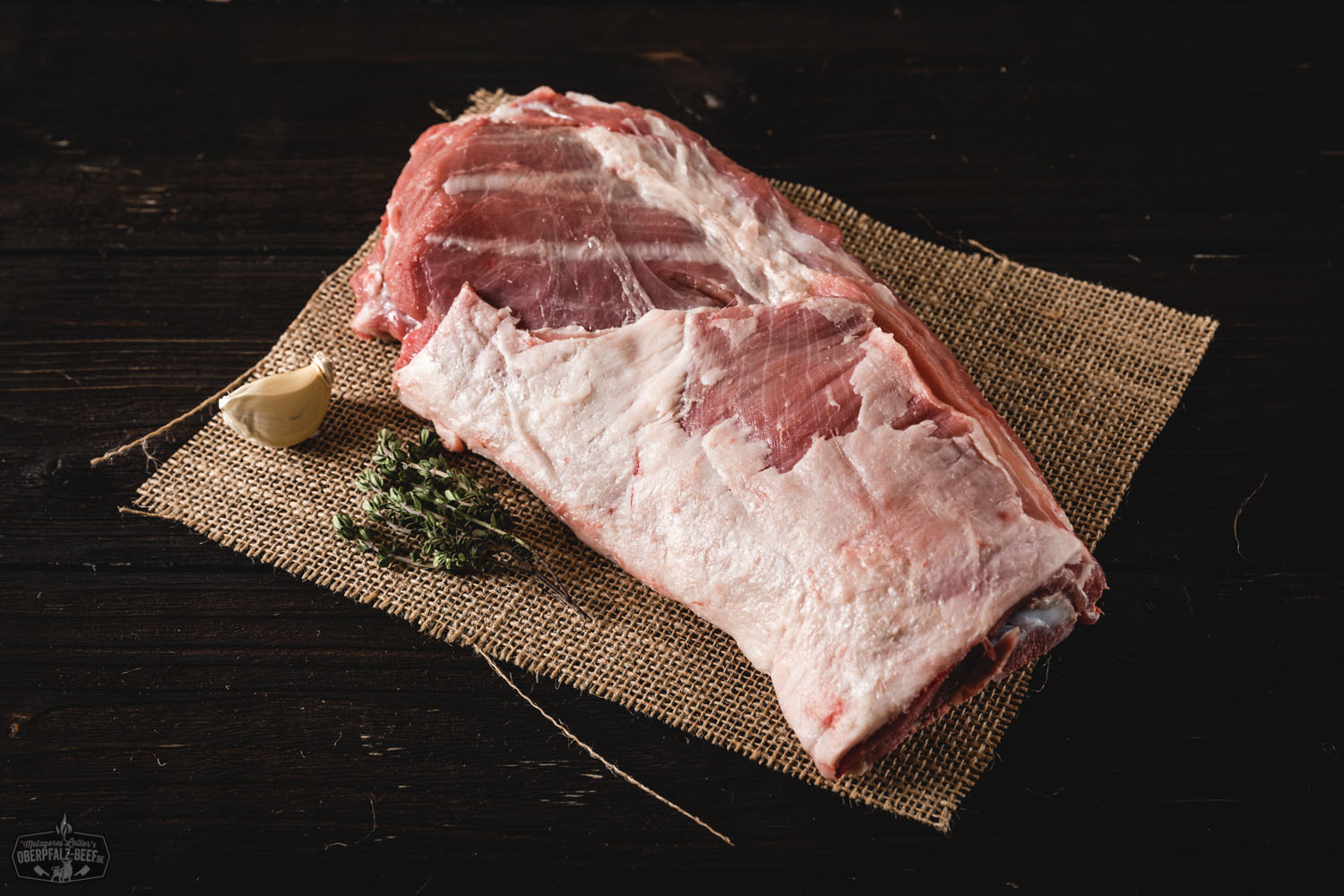 Lammhals mit Knochen vom Juradistl Lamm – Frisches Weidefleisch aus der Oberpfalz Bild eines rohen Lammhalses mit Knochen vom Juradistl-Lamm auf einer Holzoberfläche, mit Kräutern und Gewürzen garniert.